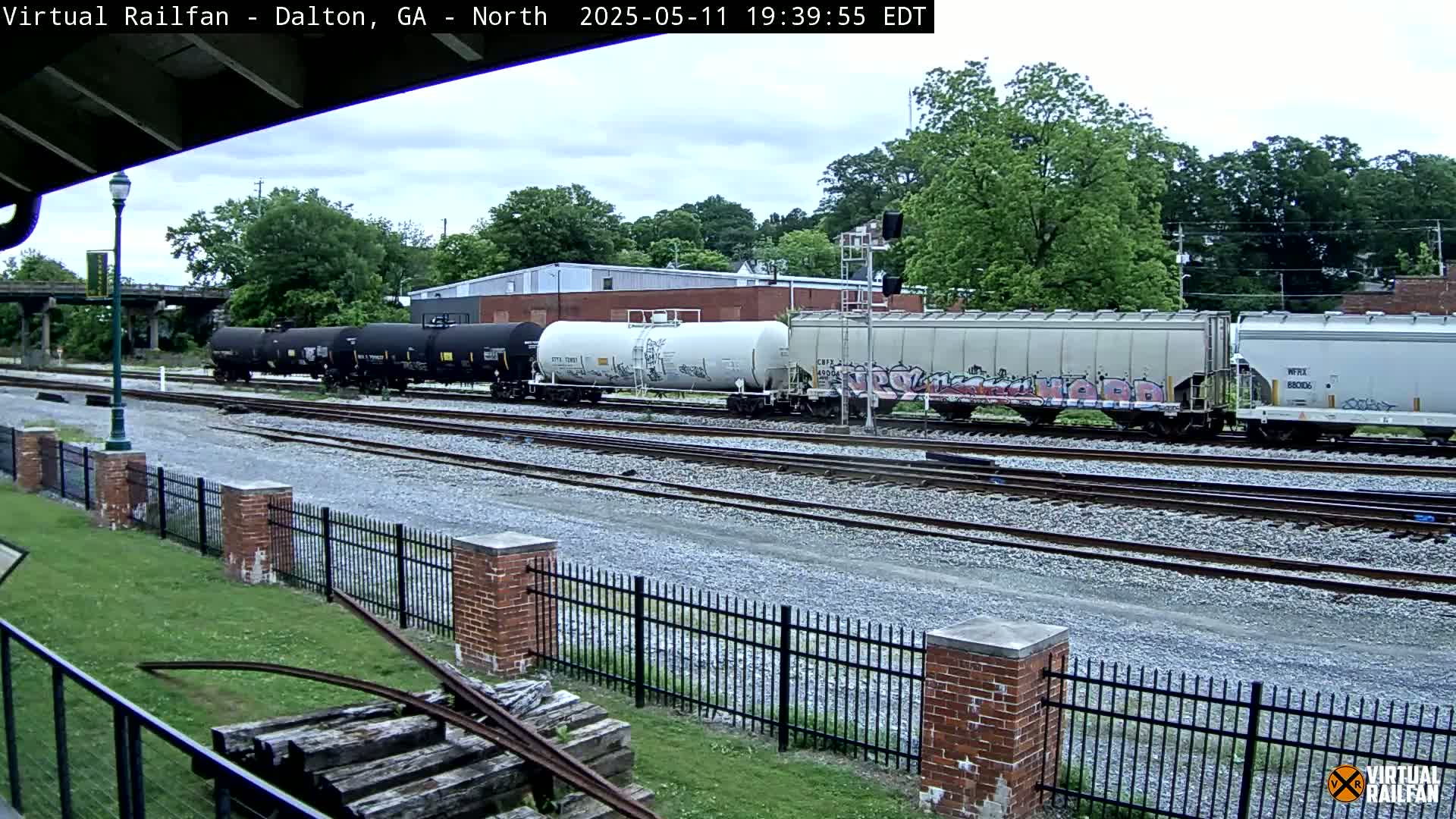 A freight train, consisting of several tanker cars and covered hopper cars, some with graffiti, sits on a rail yard under an overcast sky.