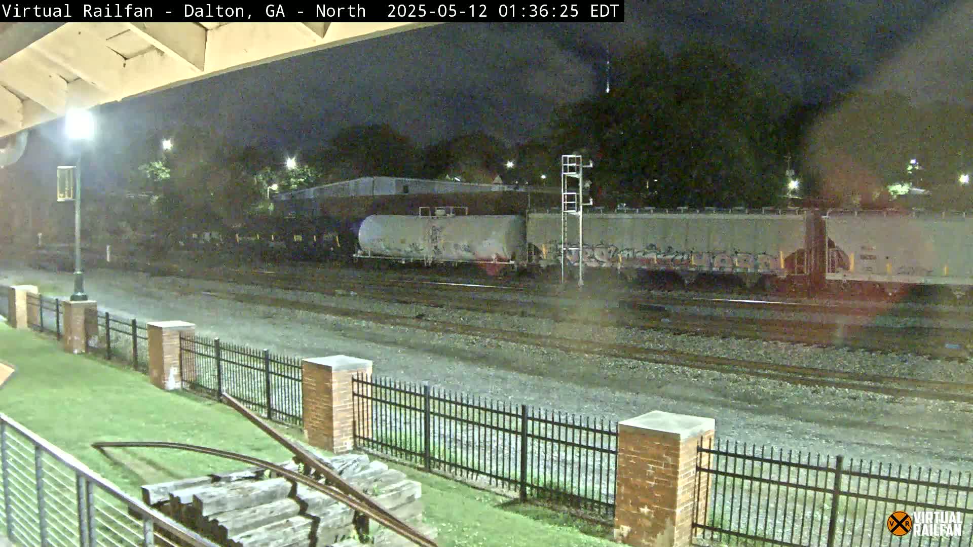 At night, a freight train with graffiti-covered cars sits on a track behind a brick fence and grassy area.