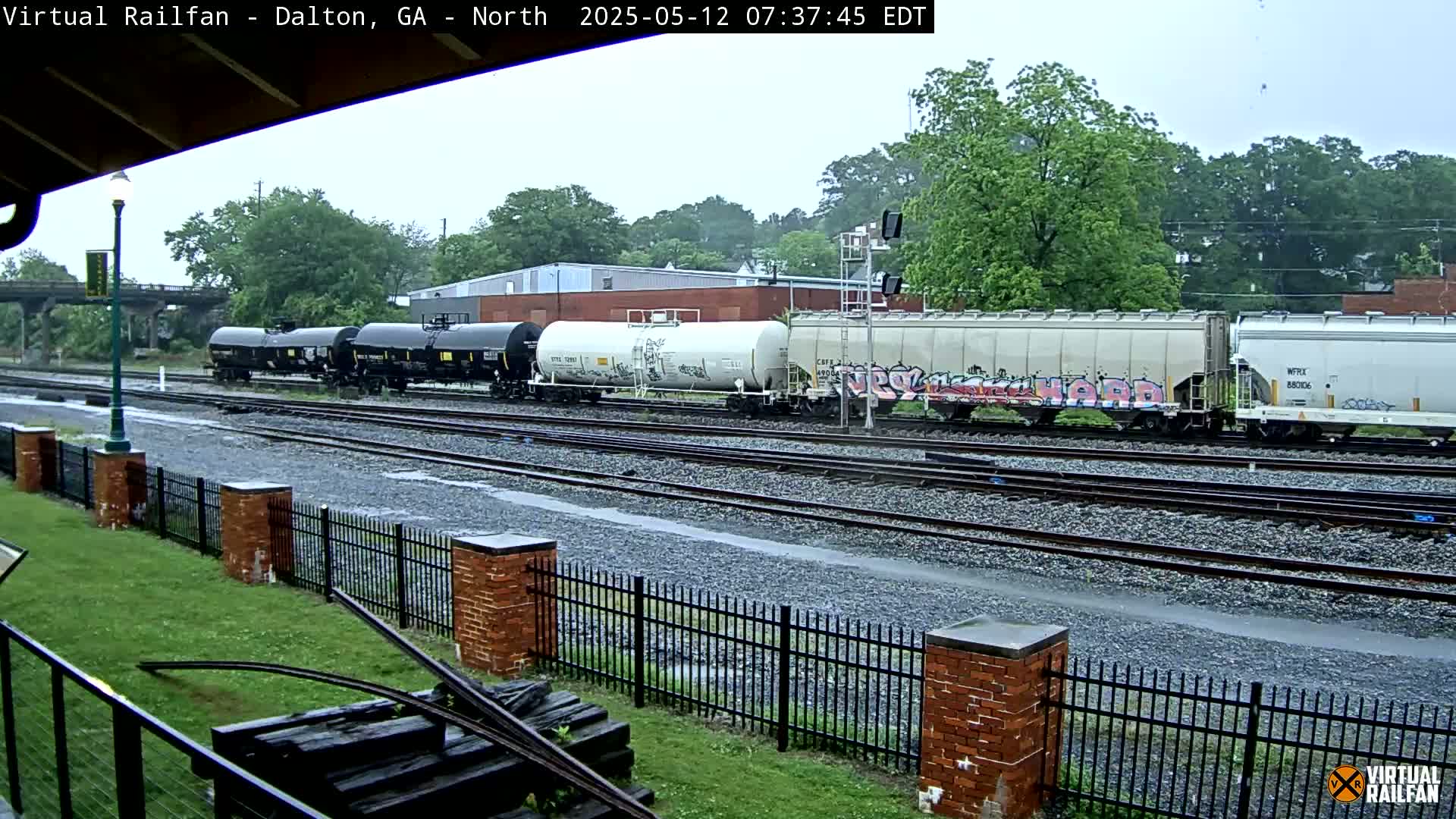 A freight train, some cars of which are graffitied, sits on a set of railway tracks in the rain, behind a short brick fence and grassy area.