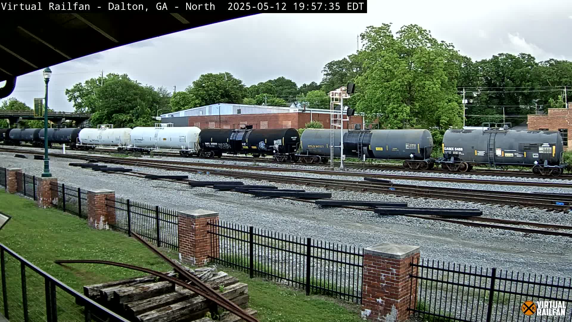 A long line of tanker cars sits on train tracks under an overcast sky next to a brick fence and grassy area.