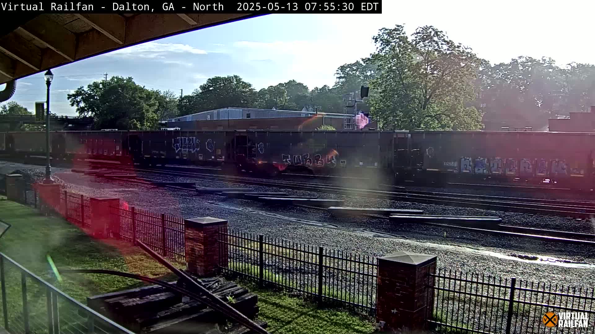 A long freight train sits on railroad tracks beside a brick fence on a sunny day.