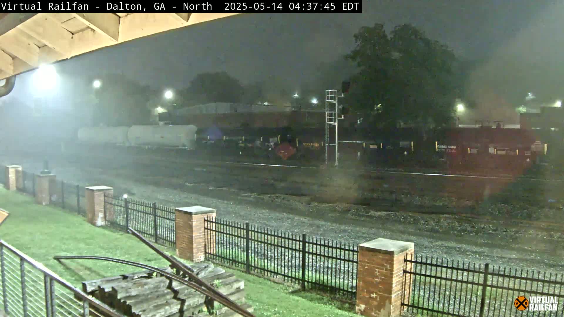 A nighttime view of a railyard during a heavy rainstorm shows a train stopped on the tracks behind a black metal fence.