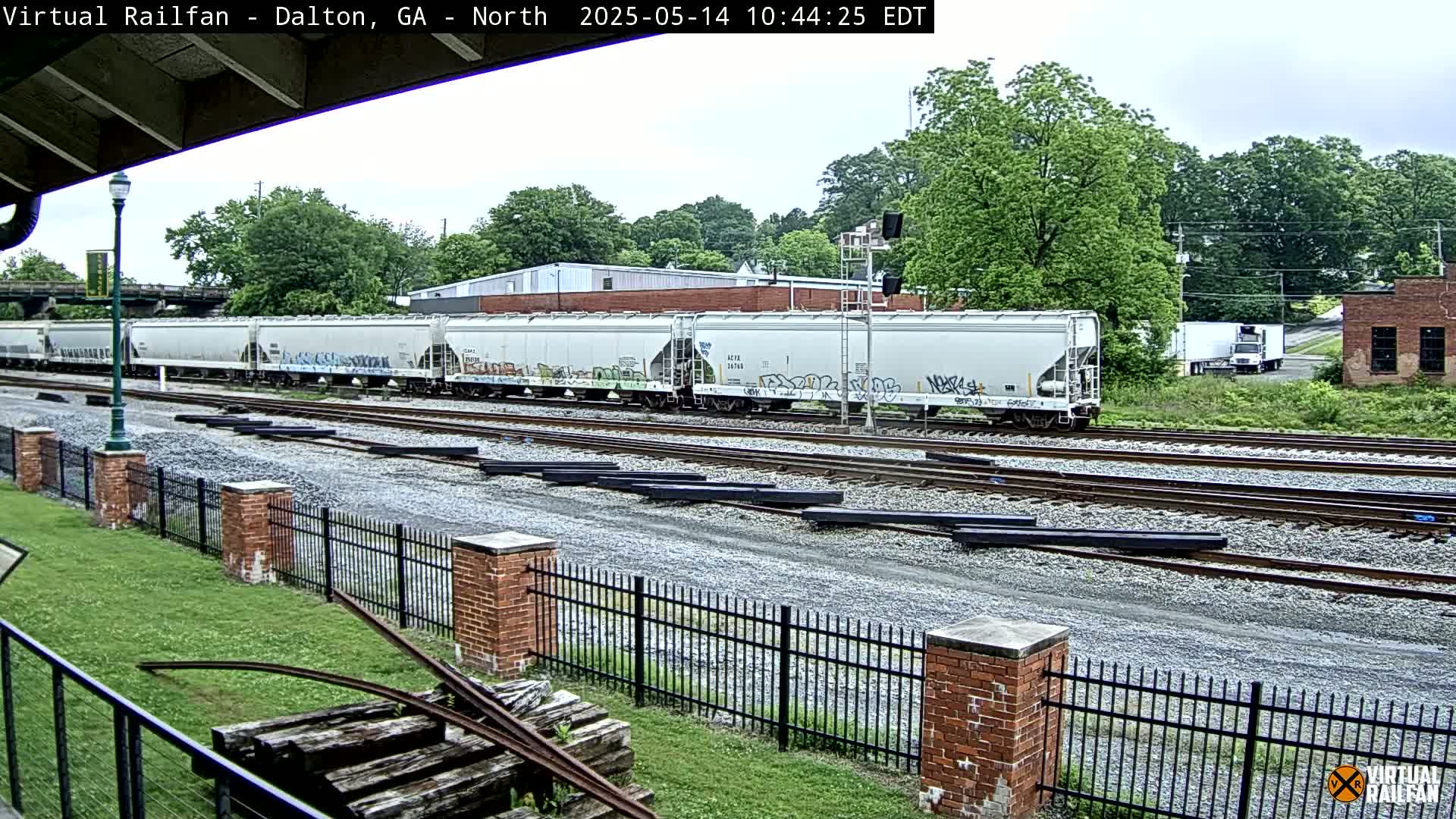 A long freight train of light-grey covered hopper cars sits on a railway track on an overcast day, beside a brick fence and green grass.
