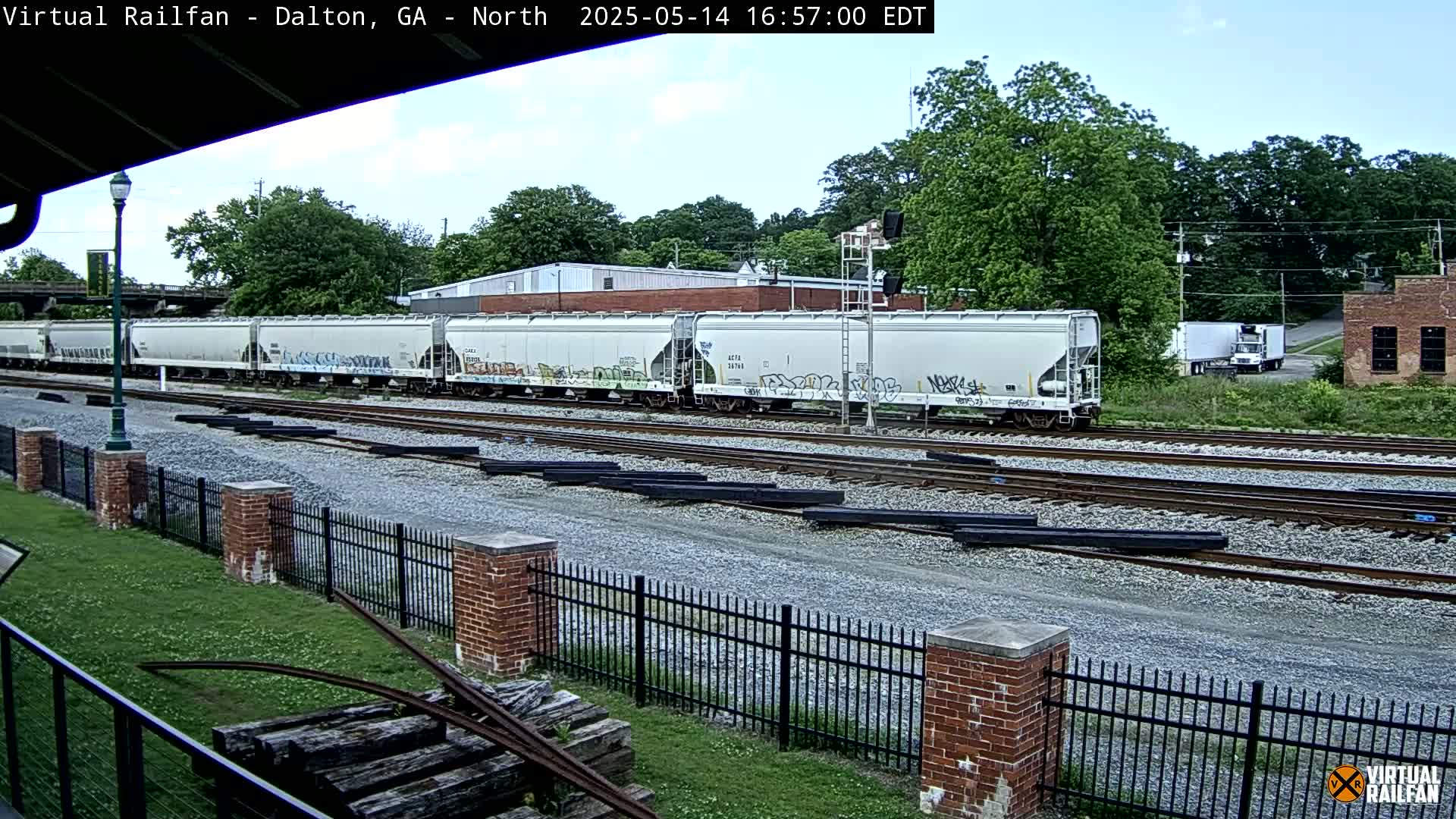 A long freight train with graffiti on its cars sits on railroad tracks next to a brick fence on an overcast day.