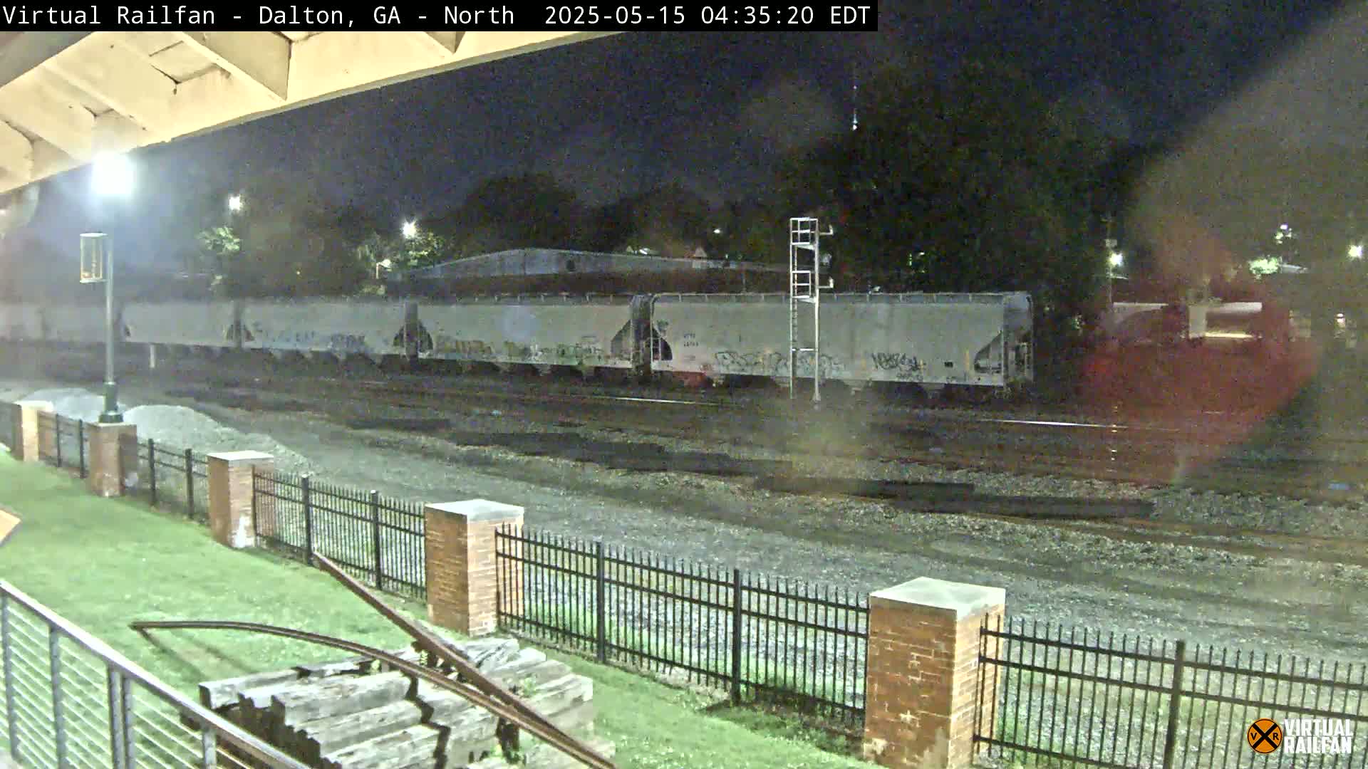 A long freight train with graffiti on its cars sits stationary at night on railroad tracks next to a metal fence.