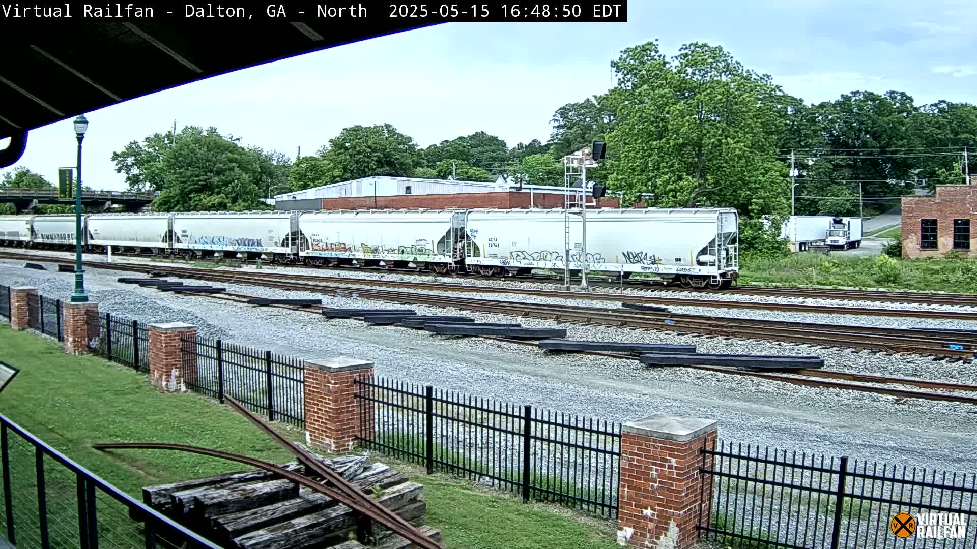 A long freight train with graffiti-covered cars sits on multiple railway tracks next to a brick fence under a partly cloudy sky.