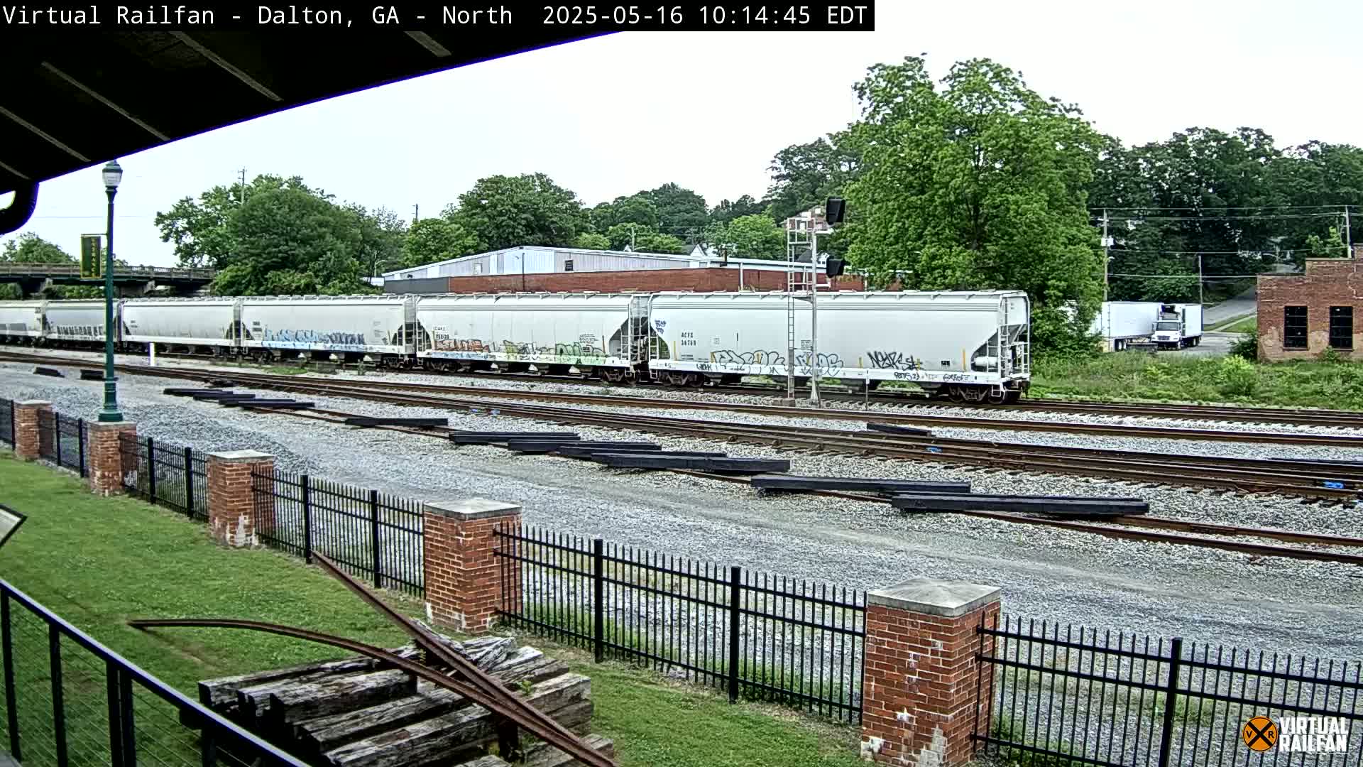 A long freight train with graffiti on its cars is seen on a railway track on an overcast day, next to a brick fence and some stacked lumber.