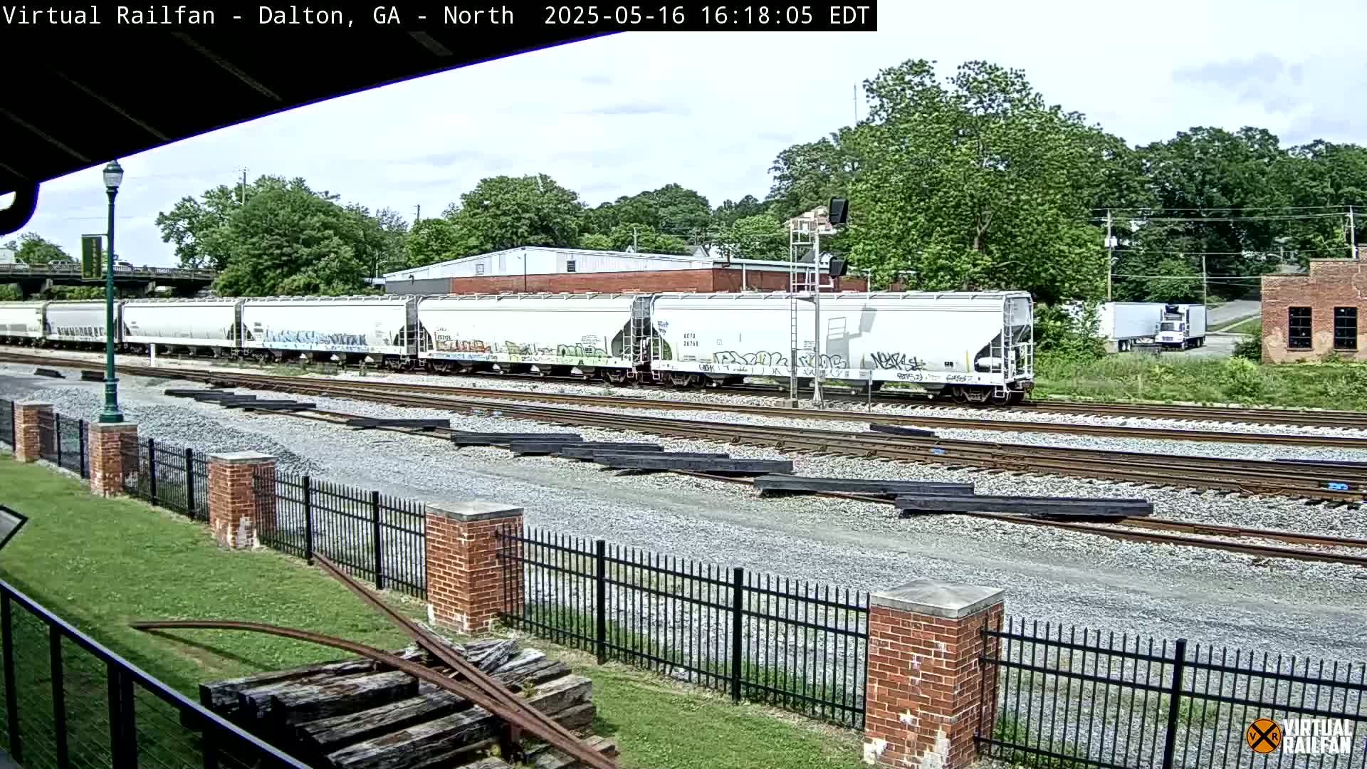 A long freight train with graffiti on some cars passes by on a sunny day, near a brick fence and a grassy area.