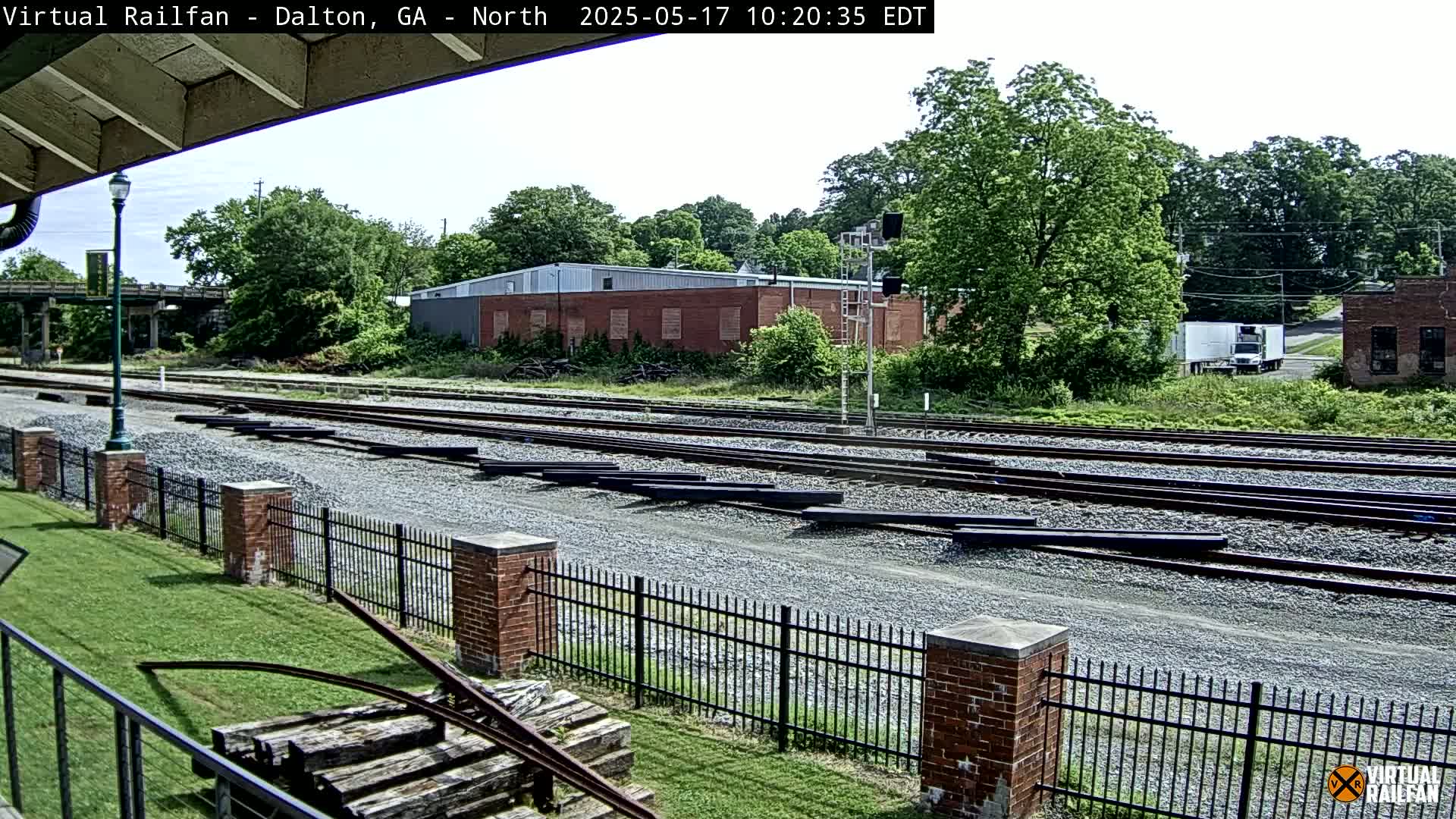 A sunny day reveals a railway yard with multiple tracks, a brick fence, and several buildings in the background, including a large brick structure and a smaller brick building.
