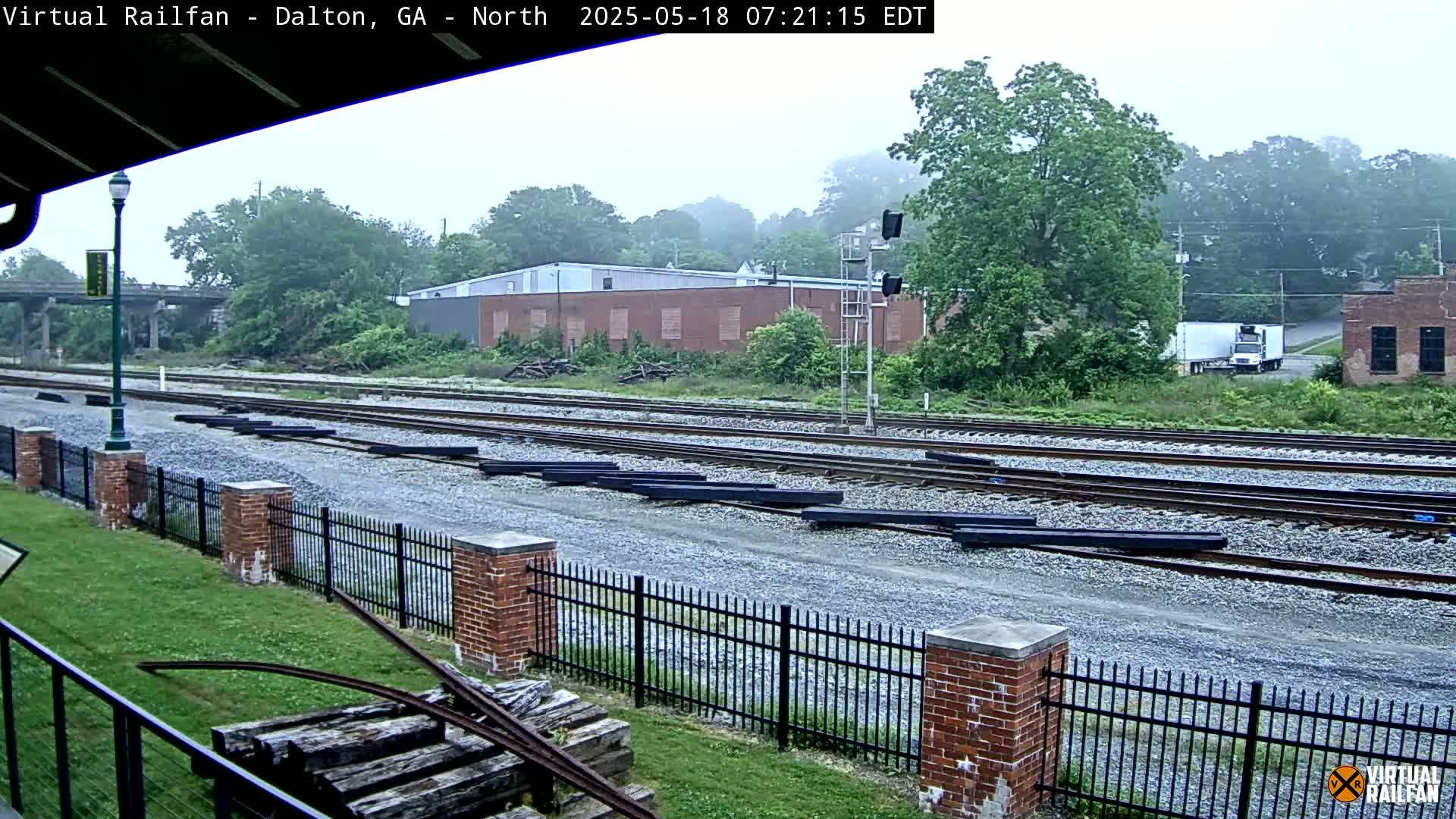 Railroad tracks extend into the distance beside a brick fence, passing industrial buildings on an overcast day.