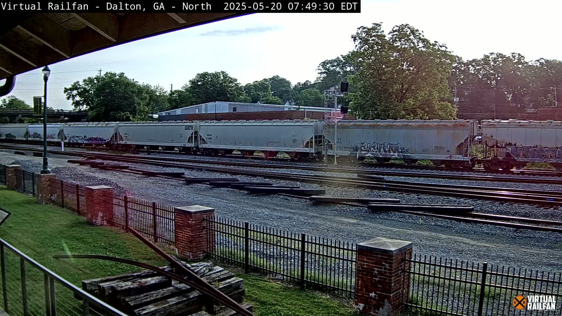 A long freight train of light gray and graffiti-covered cars sits on a set of railroad tracks under a partly cloudy sky.