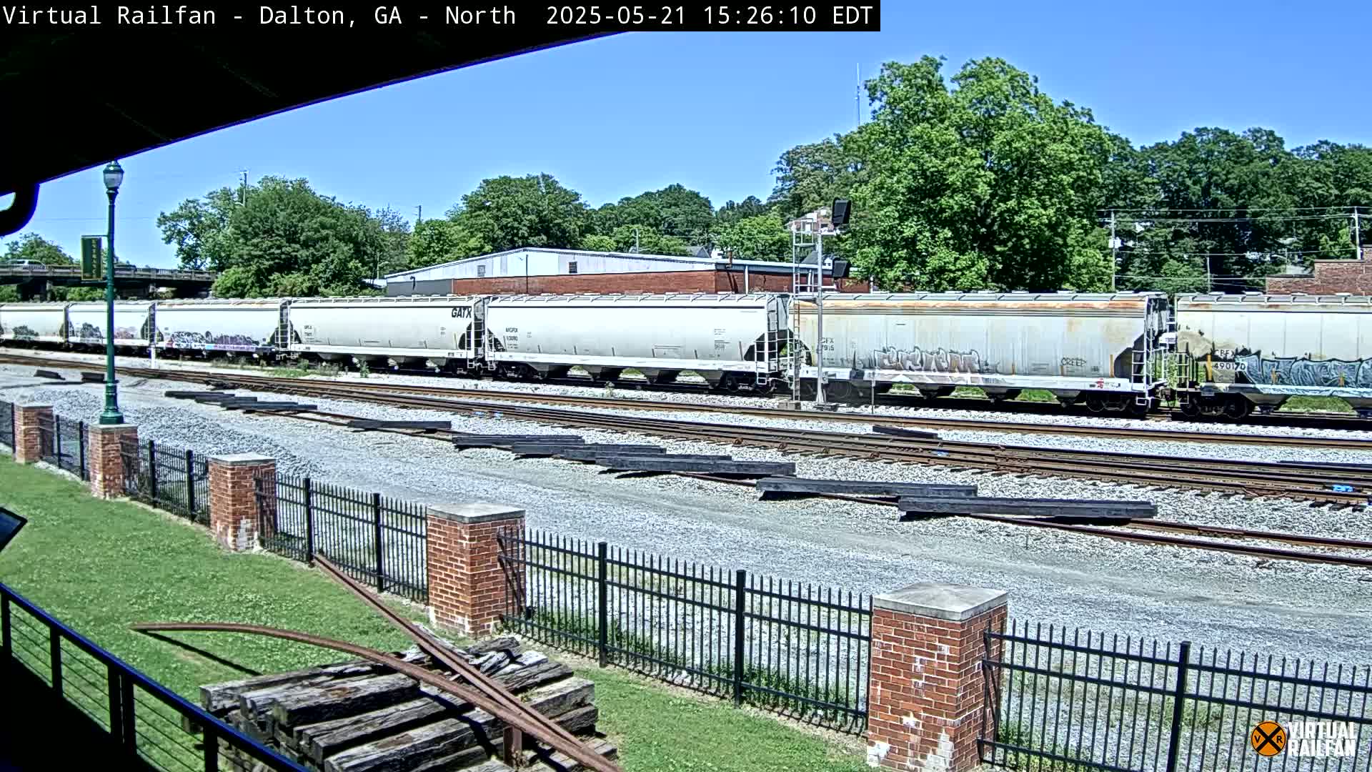 Under a sunny sky, a long freight train sits on railroad tracks near a brick fence and grassy area.