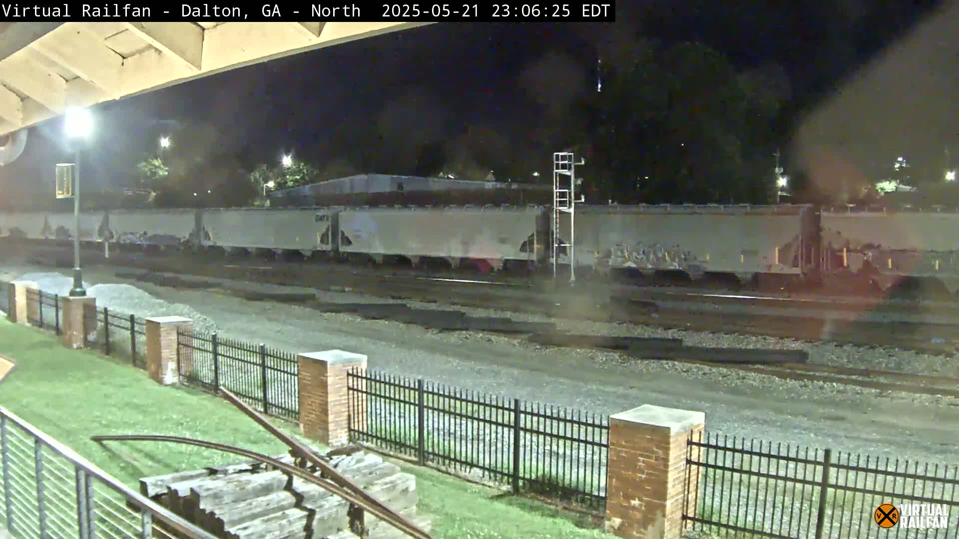 A long freight train sits on tracks at night next to a brick fence and grassy area.