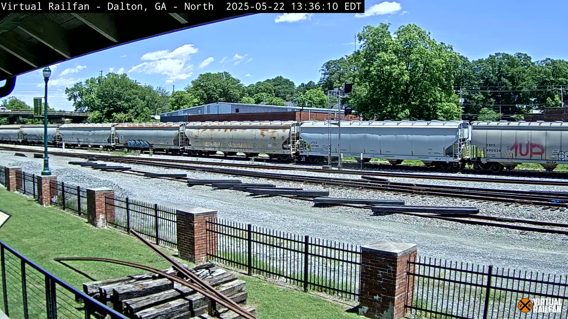 A long freight train sits on railroad tracks on a partly sunny day, next to a grassy area with a brick fence and some old wooden railroad ties.