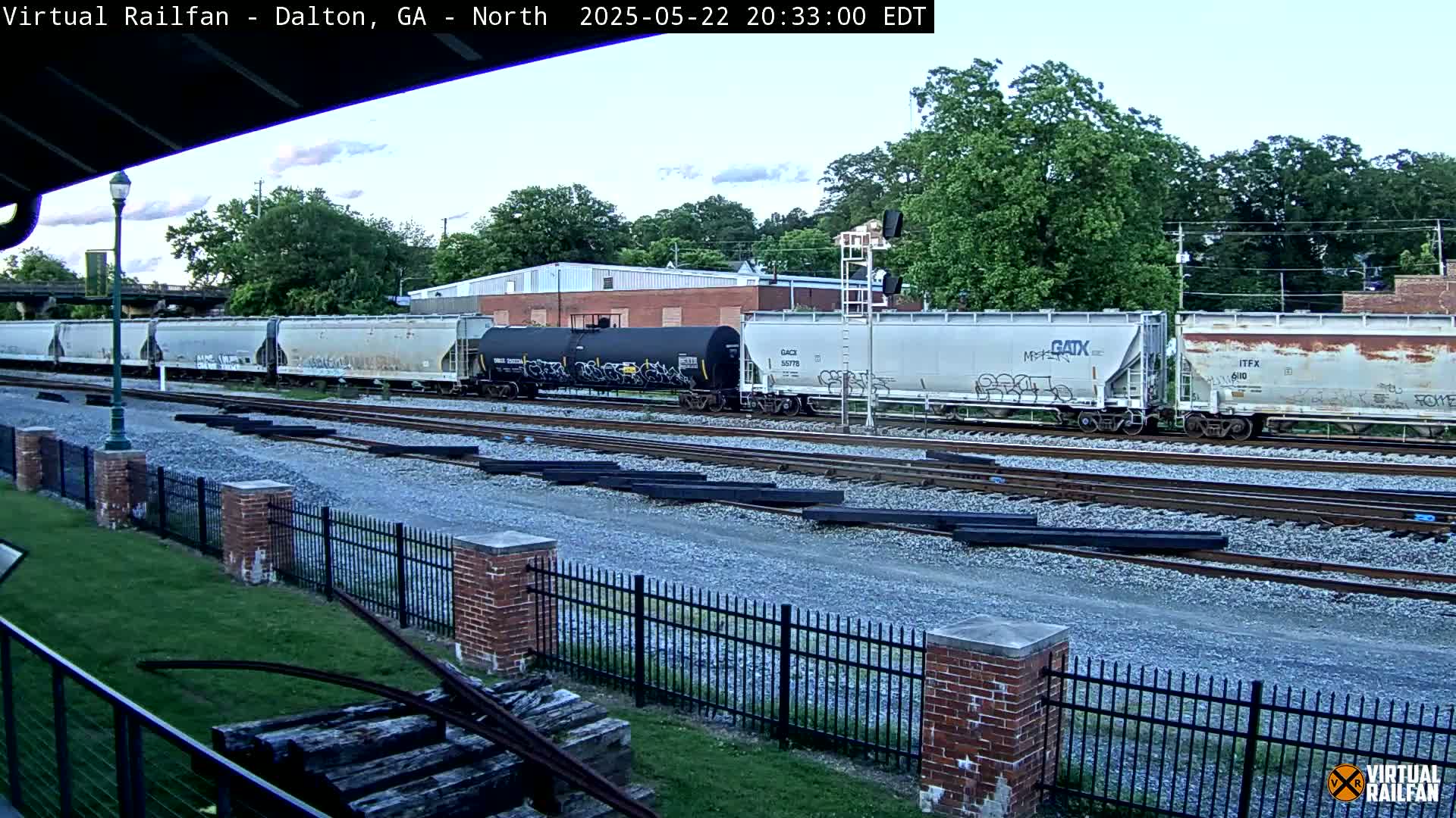A long freight train sits on multiple tracks under a partly cloudy evening sky, next to a brick fence and grassy area.