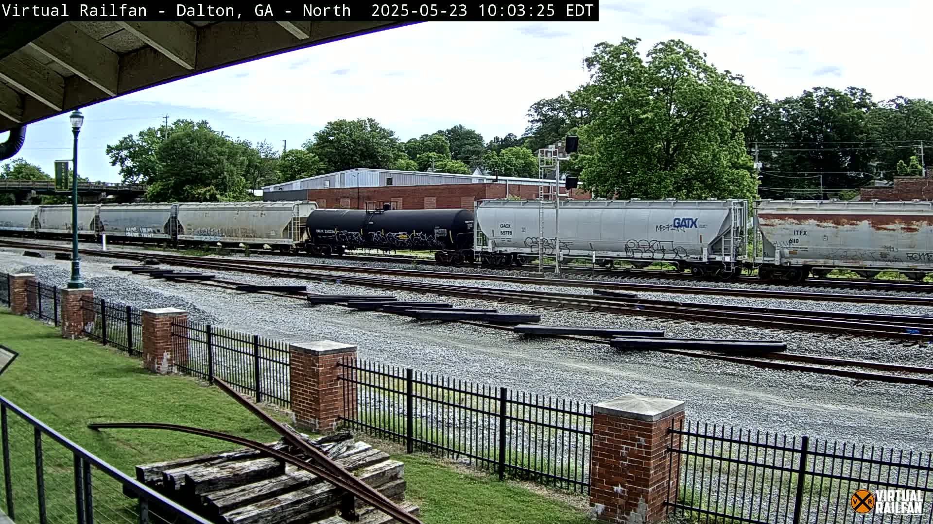 A freight train sits on a set of train tracks on an overcast day, with a brick fence and grassy area in the foreground.