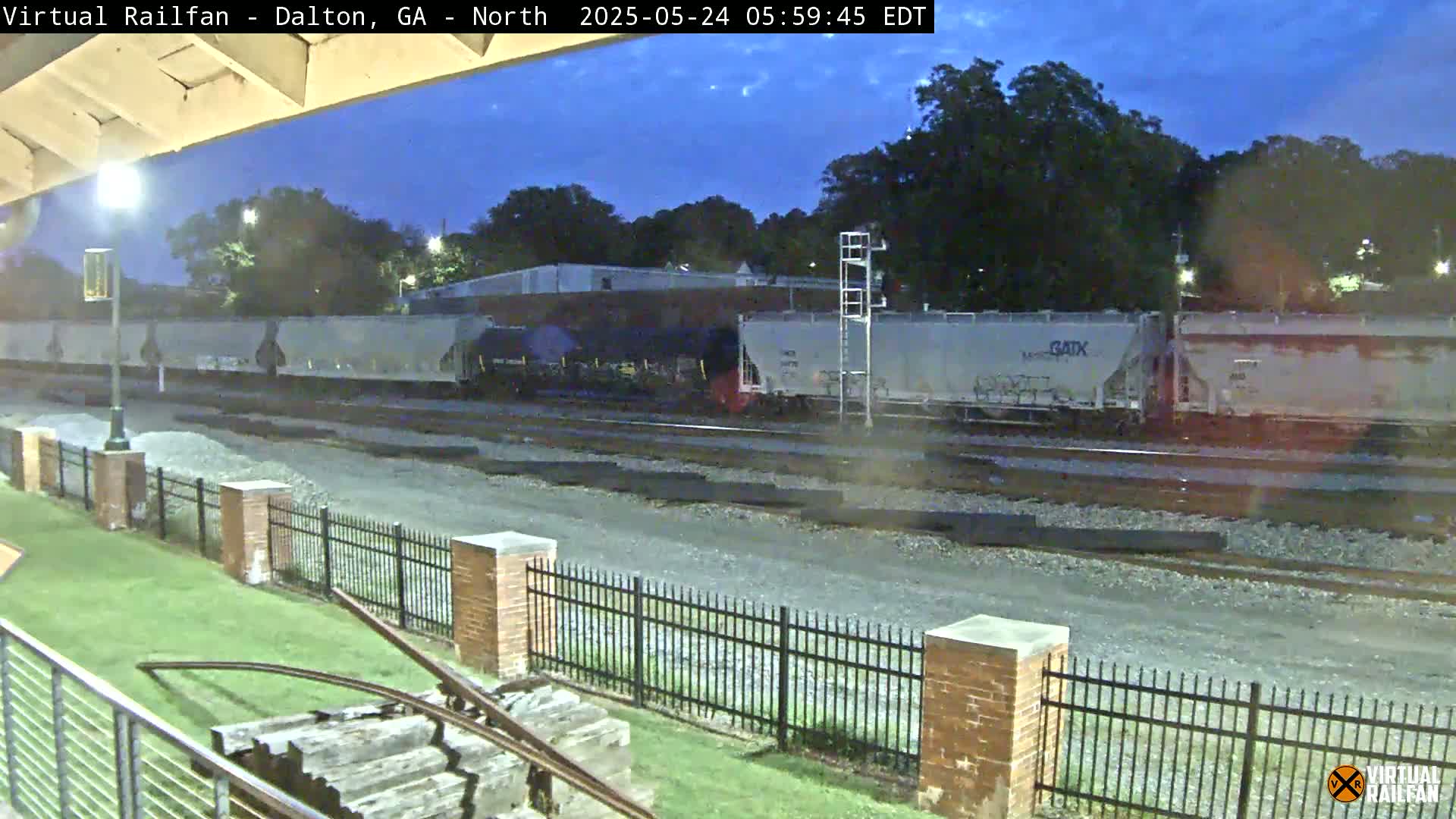 A long freight train passes by under a twilight sky next to a grassy area enclosed by a brick fence.