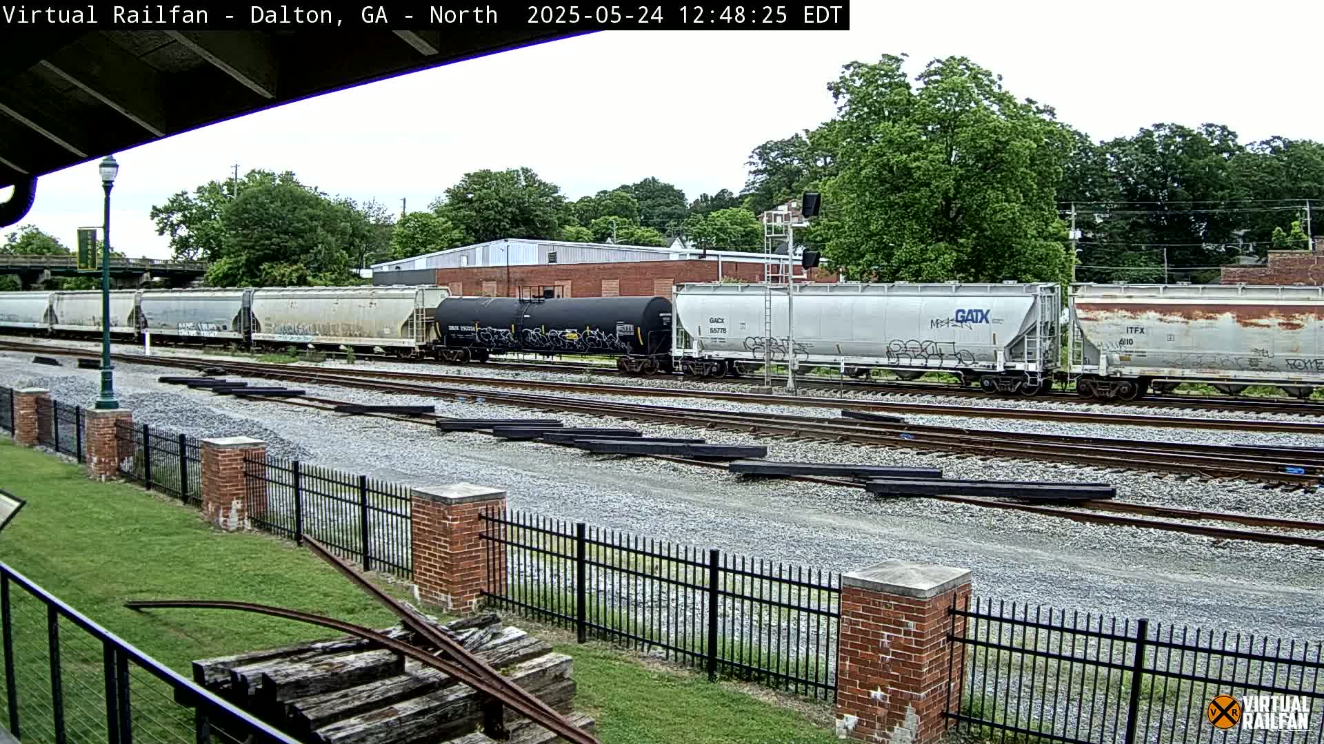A long freight train sits on multiple railway tracks next to a brick fence and grassy area under an overcast sky.