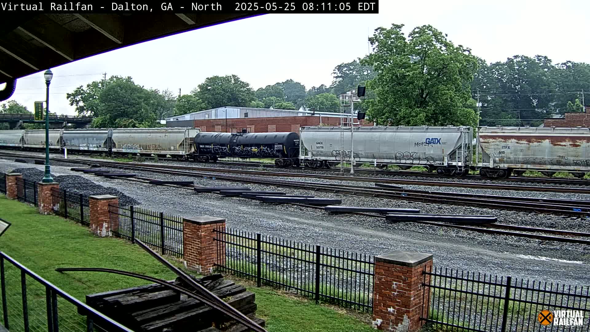 A freight train, consisting of several tanker and hopper cars, sits on multiple railway tracks in front of a brick building under an overcast sky.
