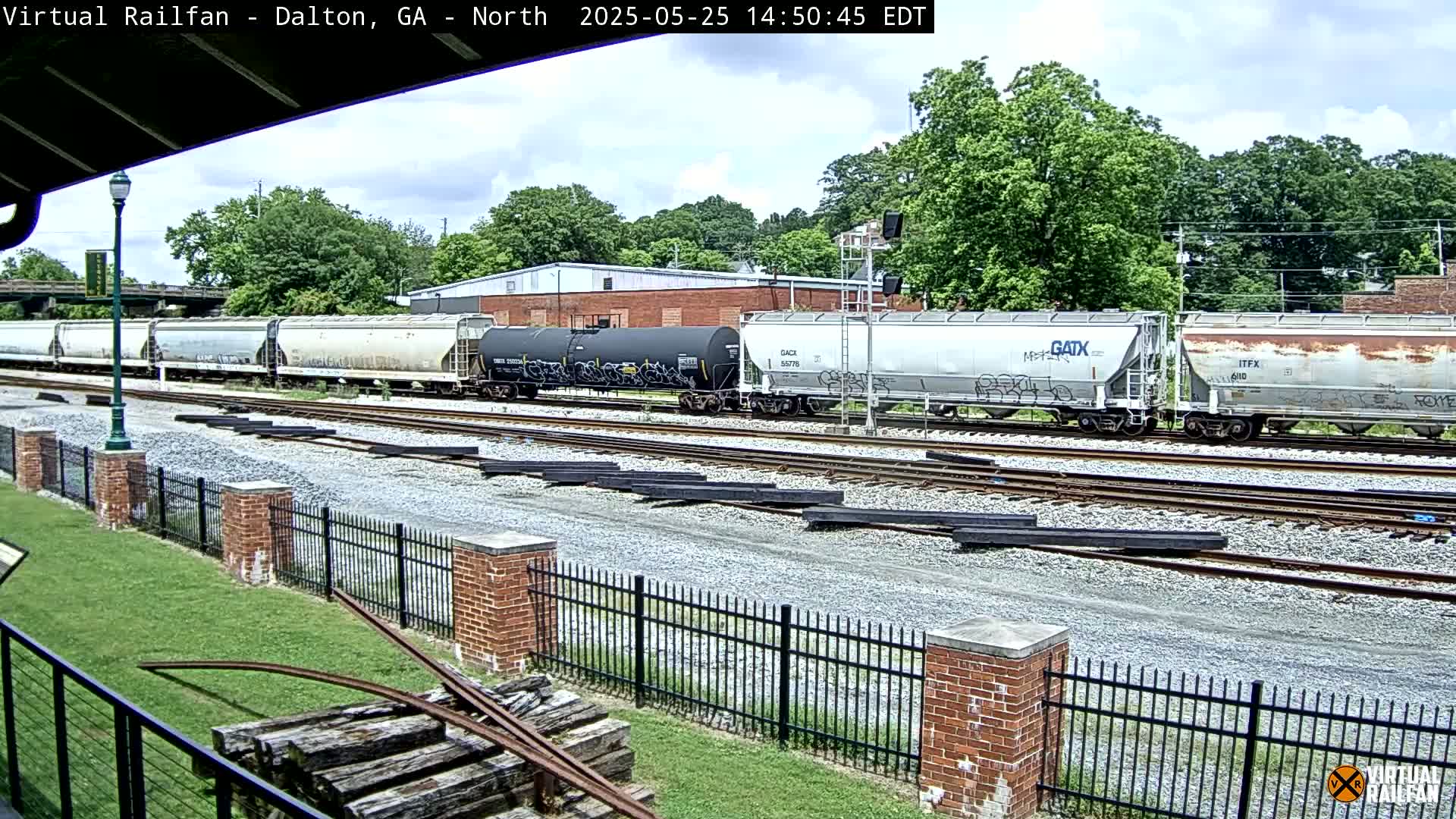 A long freight train sits on railroad tracks under a partly cloudy sky, next to a brick fence and grassy area.
