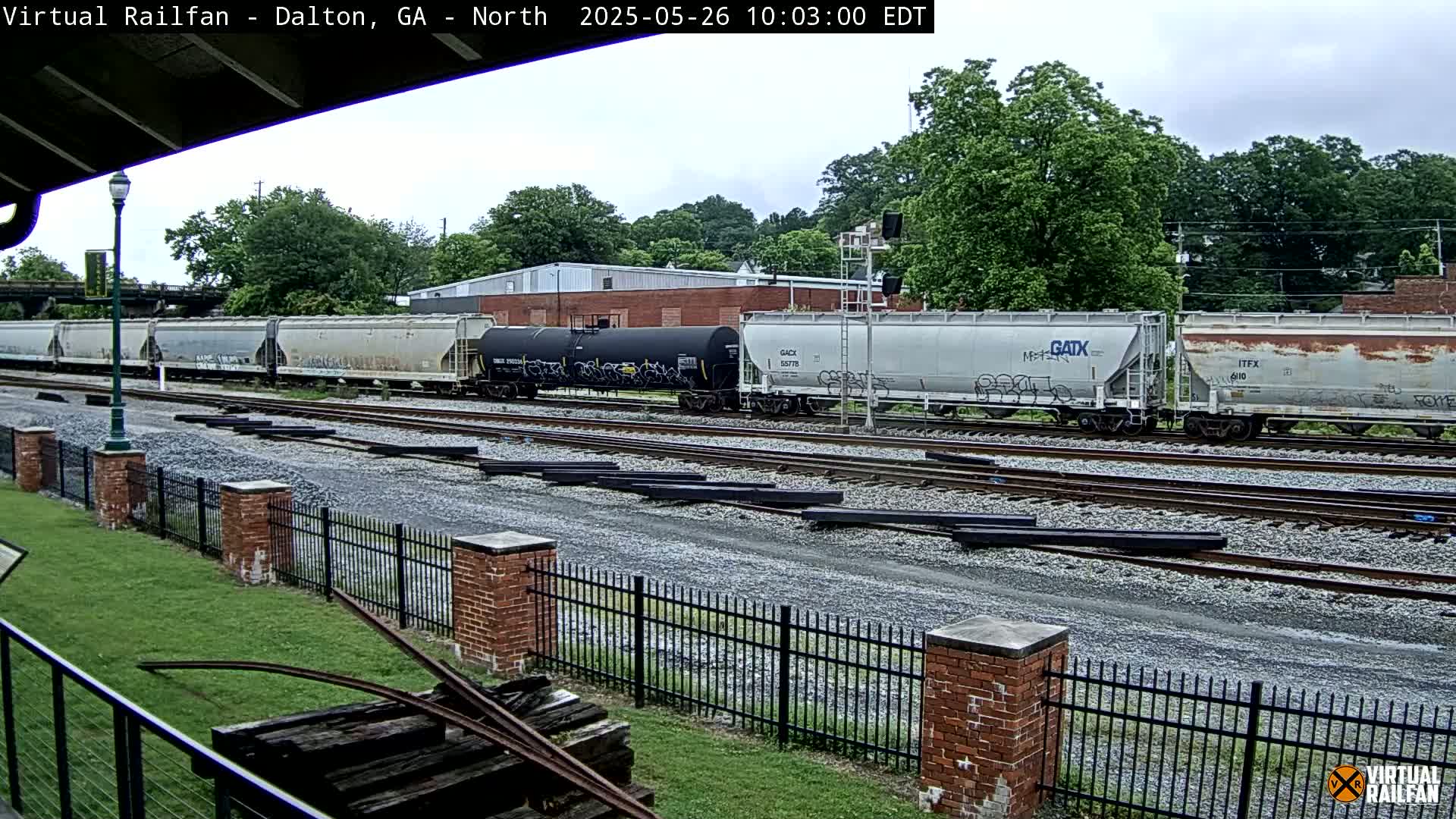 A freight train with multiple cars sits on railway tracks beside a grassy area enclosed by a brick fence, under an overcast sky.