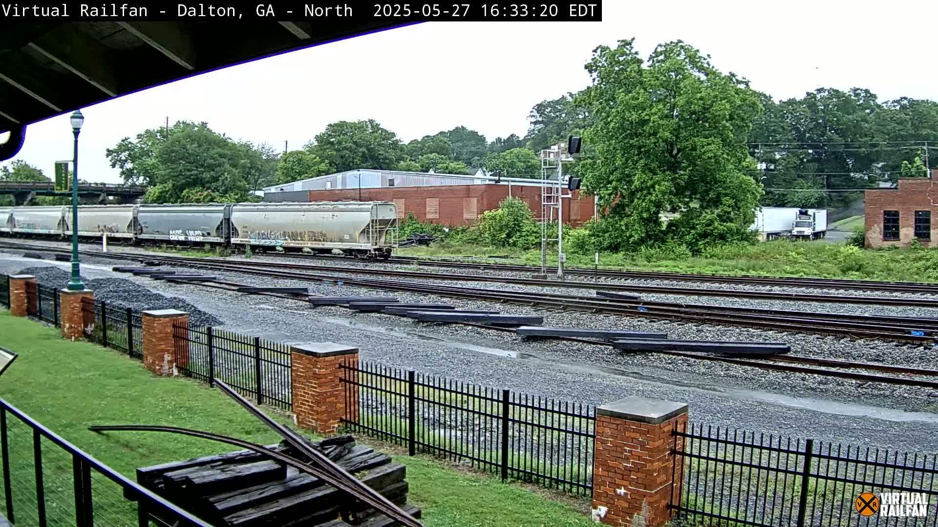 A freight train sits on railroad tracks next to a brick building on a rainy day, with a black metal fence separating the tracks from a grassy area.