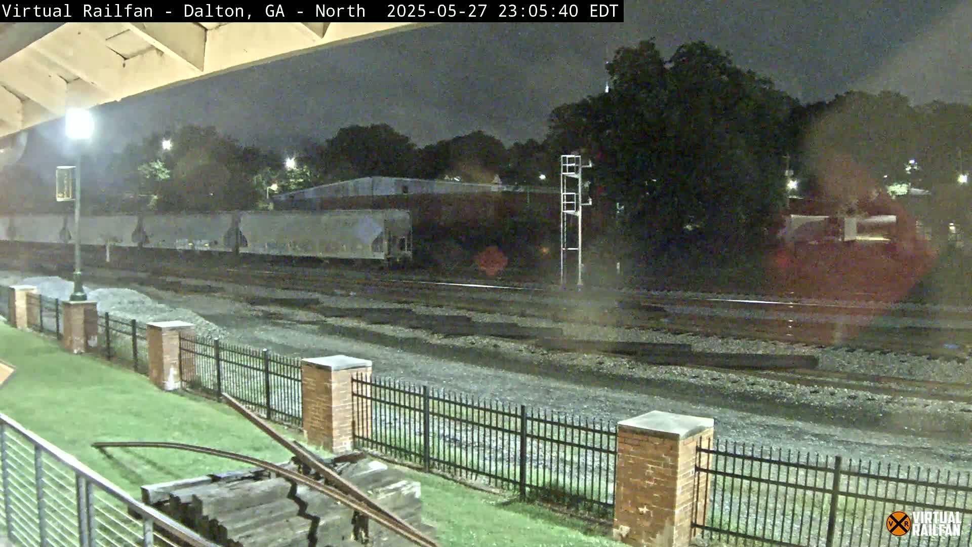 A nighttime view of railroad tracks with a freight train in the distance, under a dark sky, behind a short brick fence and grassy area.