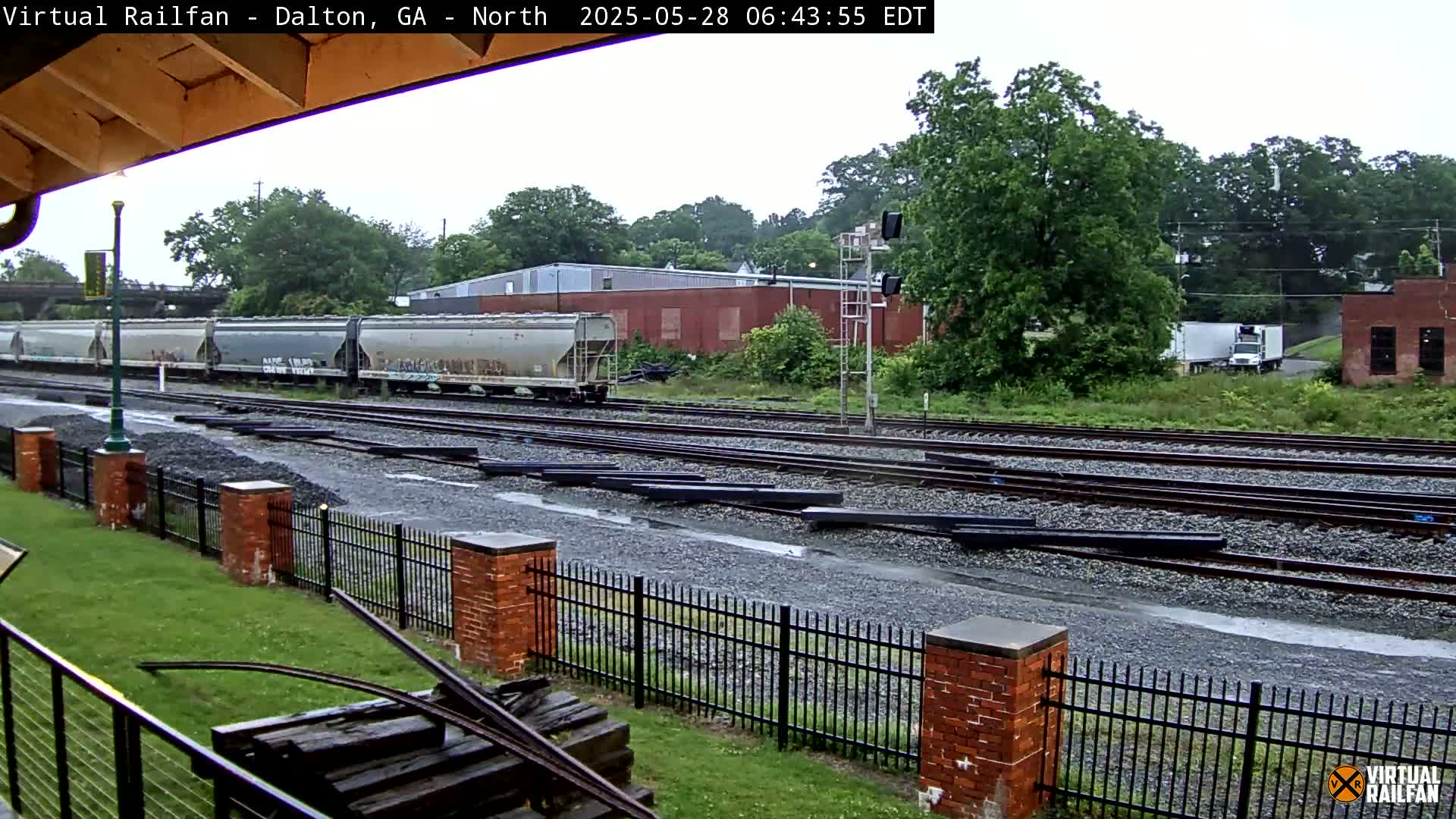 A rain-soaked railway yard with several railcars, a brick fence, and a distant industrial building is seen.