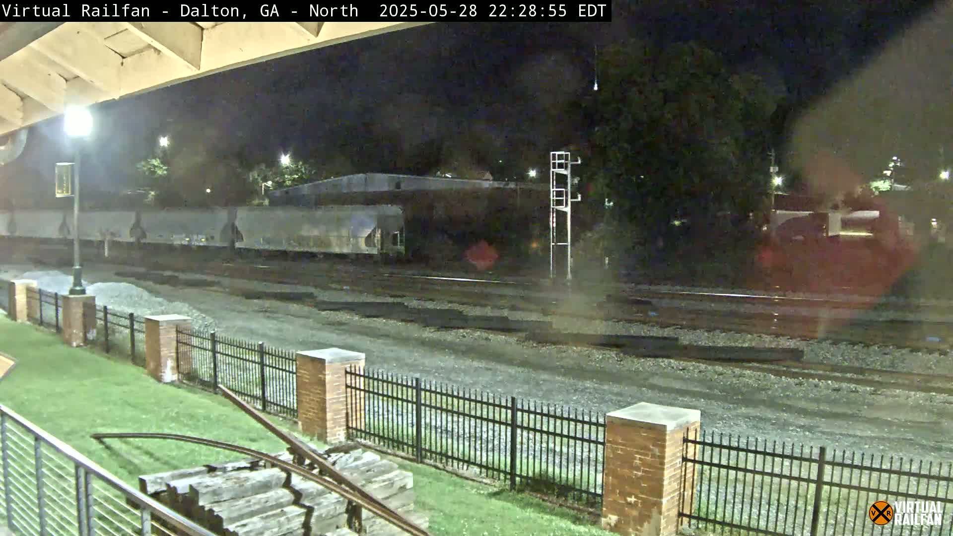 A nighttime view of railroad tracks and a train, with a metal fence and a grassy area in the foreground, under a clear night sky.