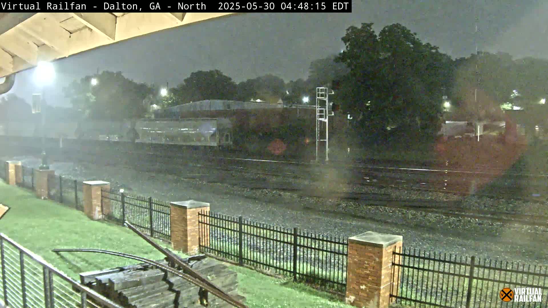 A nighttime view of a rain-soaked railway track behind a brick fence, with a train car visible in the background under heavy rain.