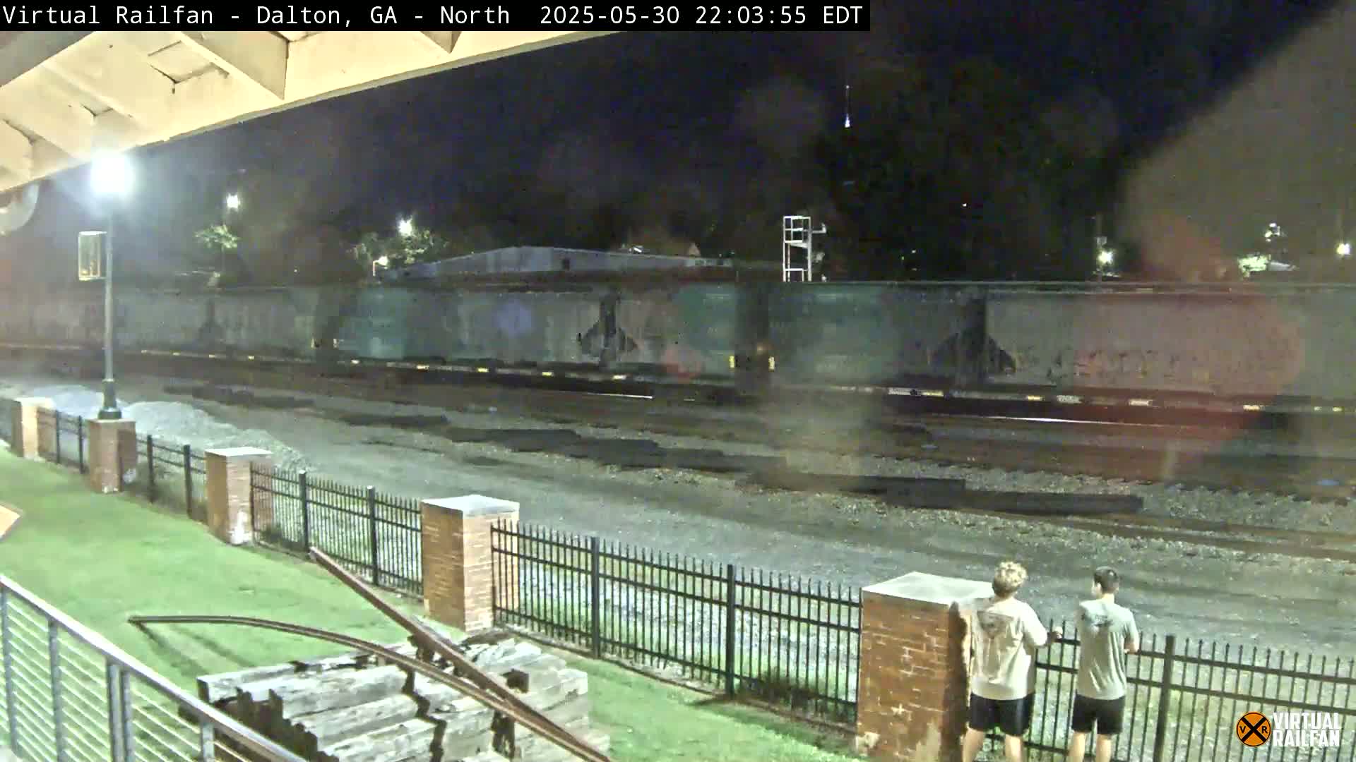 At night, a long freight train passes by a fenced-in area where two people stand watching, under a bridge.