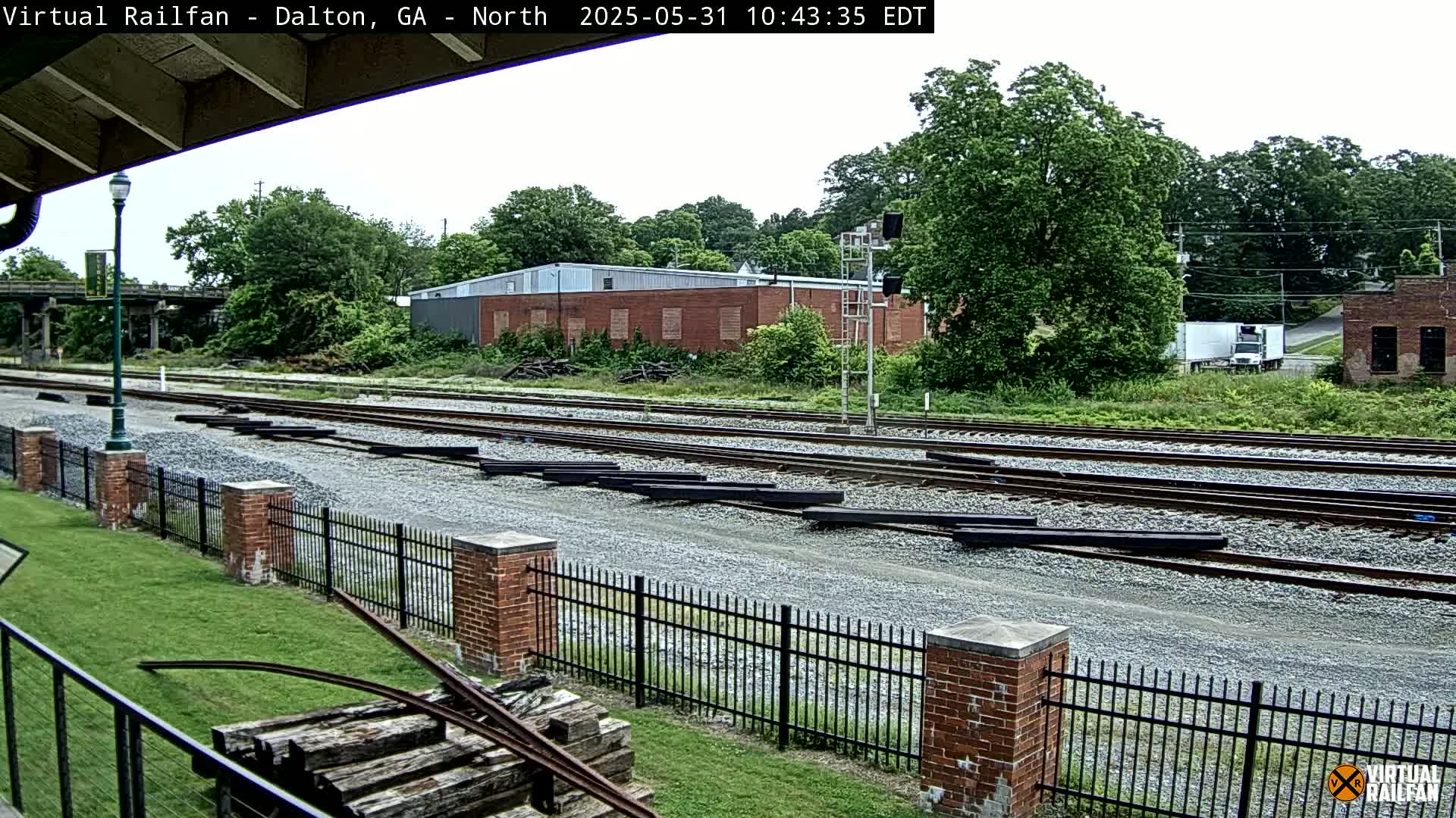 A view of railroad tracks and a brick building on a partly cloudy day, with green trees and grass surrounding the area.