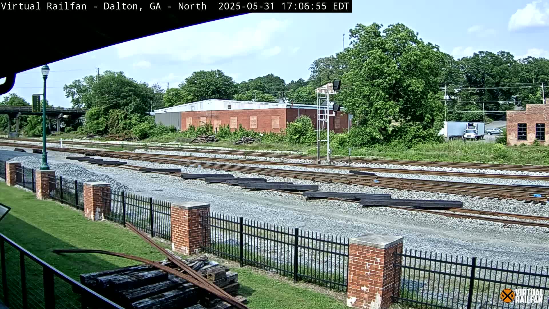 A sunny day view of railroad tracks with several long pieces of metal stacked near a brick fence and some buildings in the background.