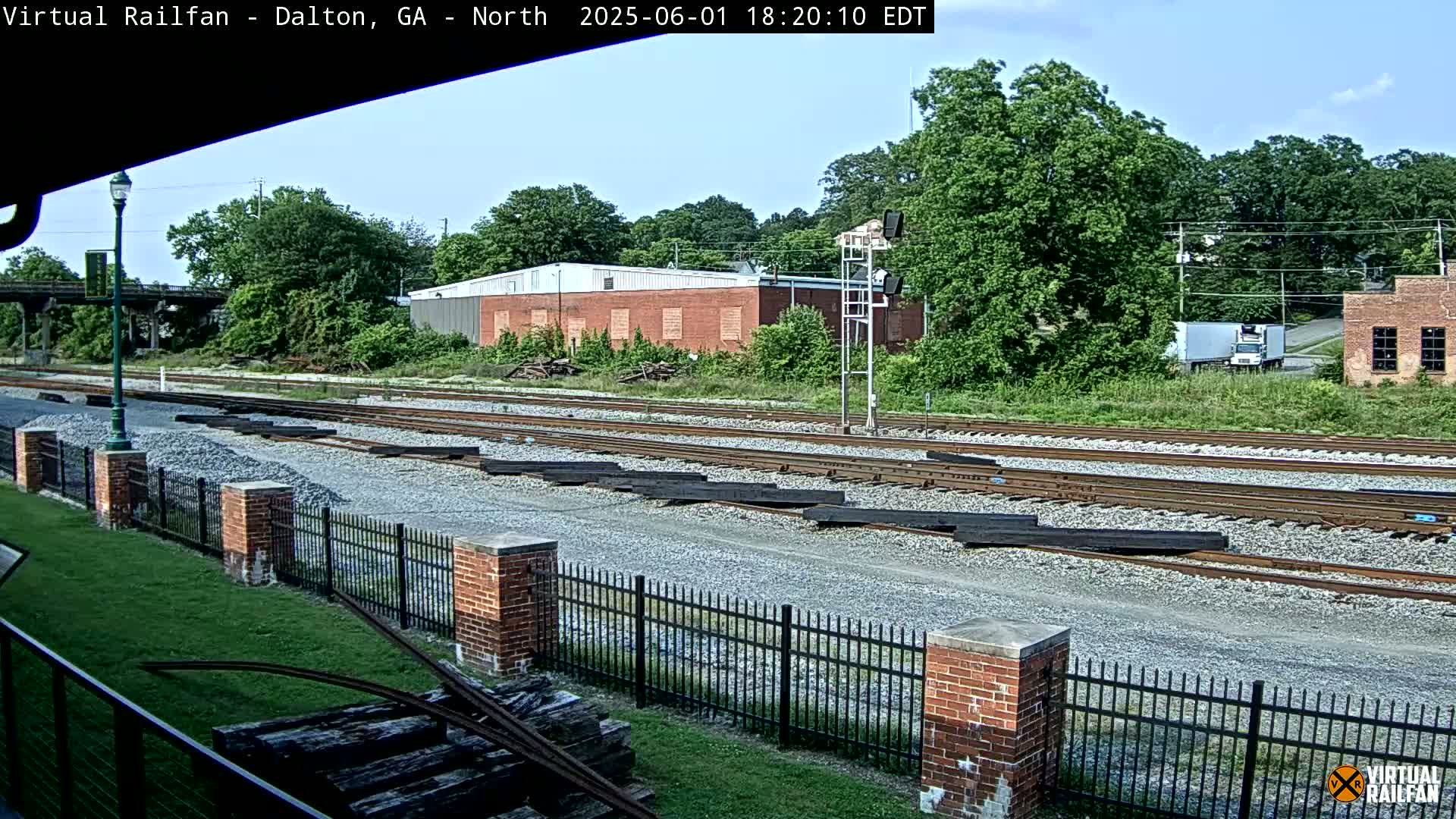 A sunny day view of railroad tracks with stacks of lumber beside them, a brick building in the background, and lush green trees.