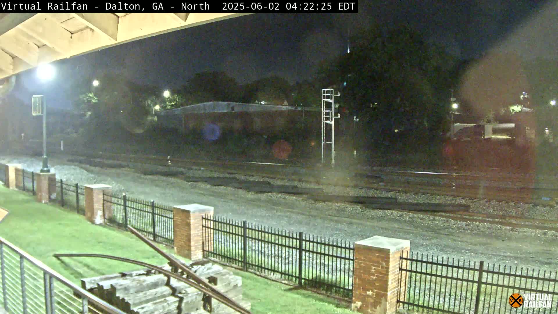 A nighttime view of railroad tracks, bordered by a black metal fence and brick pillars, with a grassy area and part of a wooden structure visible in the foreground under a clear night sky.