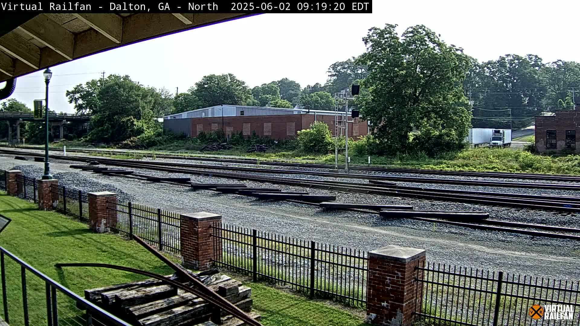 A sunny view of railroad tracks and switching yards, bordered by a brick fence and lush green grass, with buildings and trees in the background.