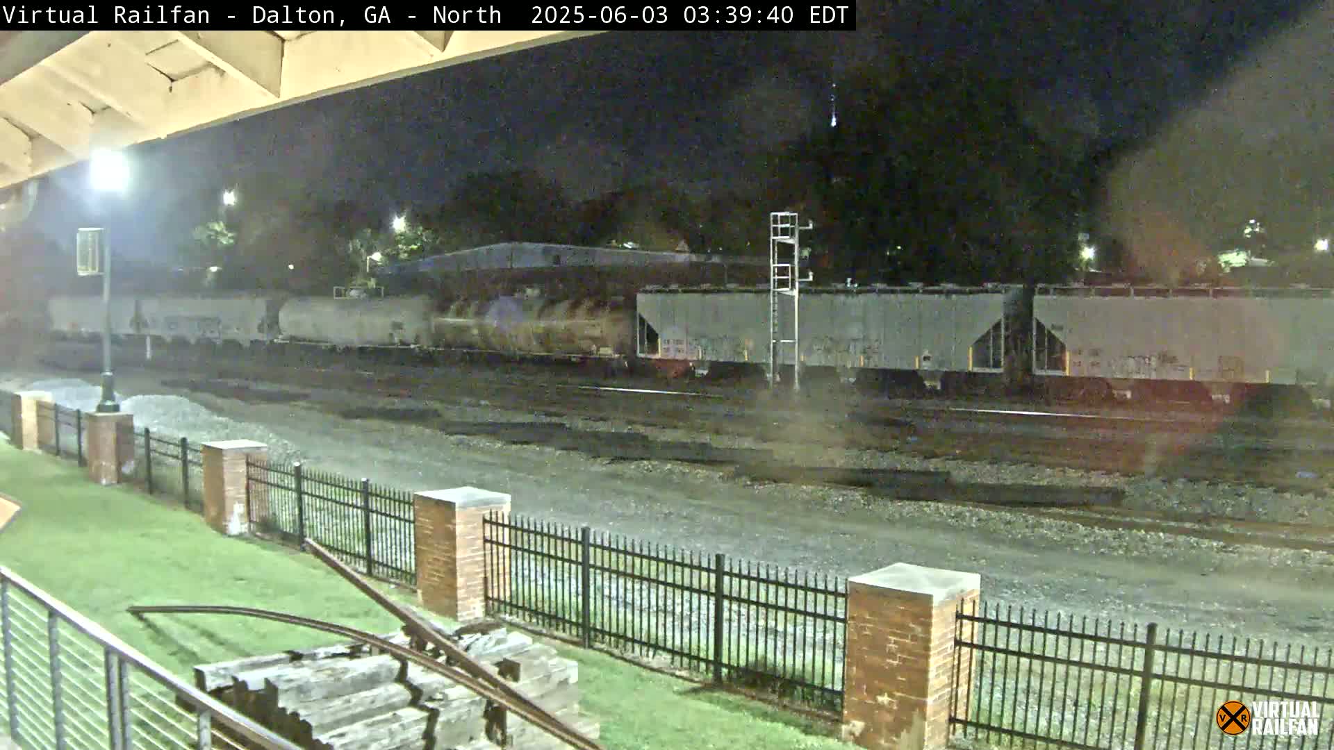 A long freight train passes by at night under a mostly clear sky, next to a metal fence and grassy area.