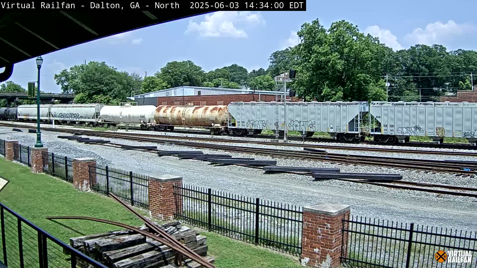 A freight train with several cars, some showing graffiti, sits on railroad tracks under a partly cloudy sky next to a brick fence and lush green grass.