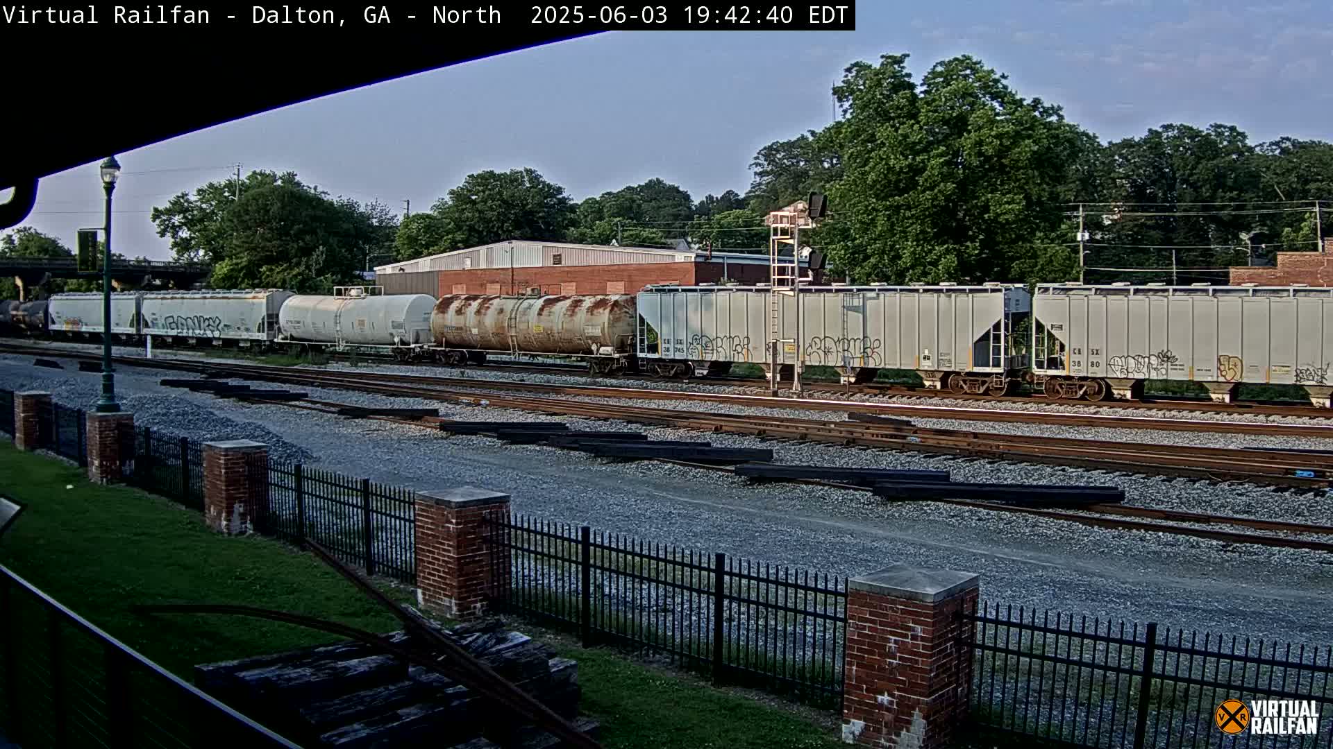 Under a clear sky, several freight cars sit on a railway track next to a brick fence, with trees and industrial buildings in the background.