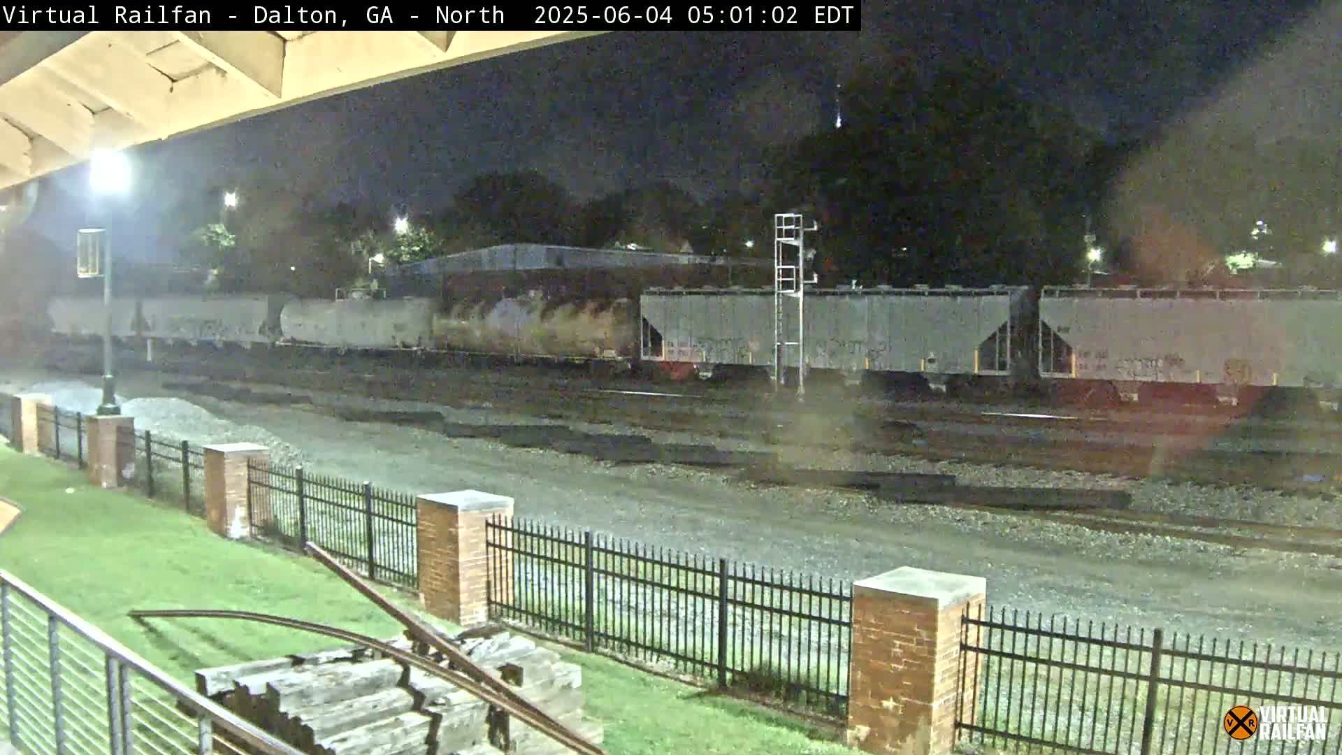 At night, a long freight train sits stationary on tracks behind a brick fence and grassy area.