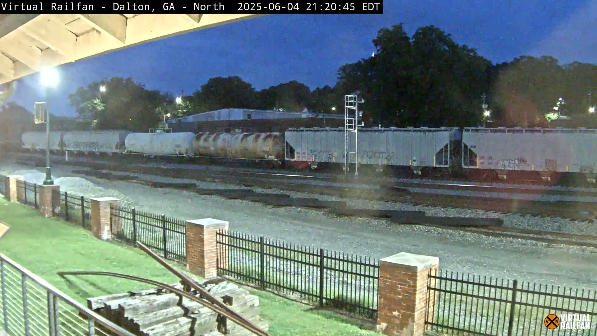 Under a twilight sky, a freight train consisting of several boxcars and tank cars sits on railway tracks near a brick fence and grassy area.