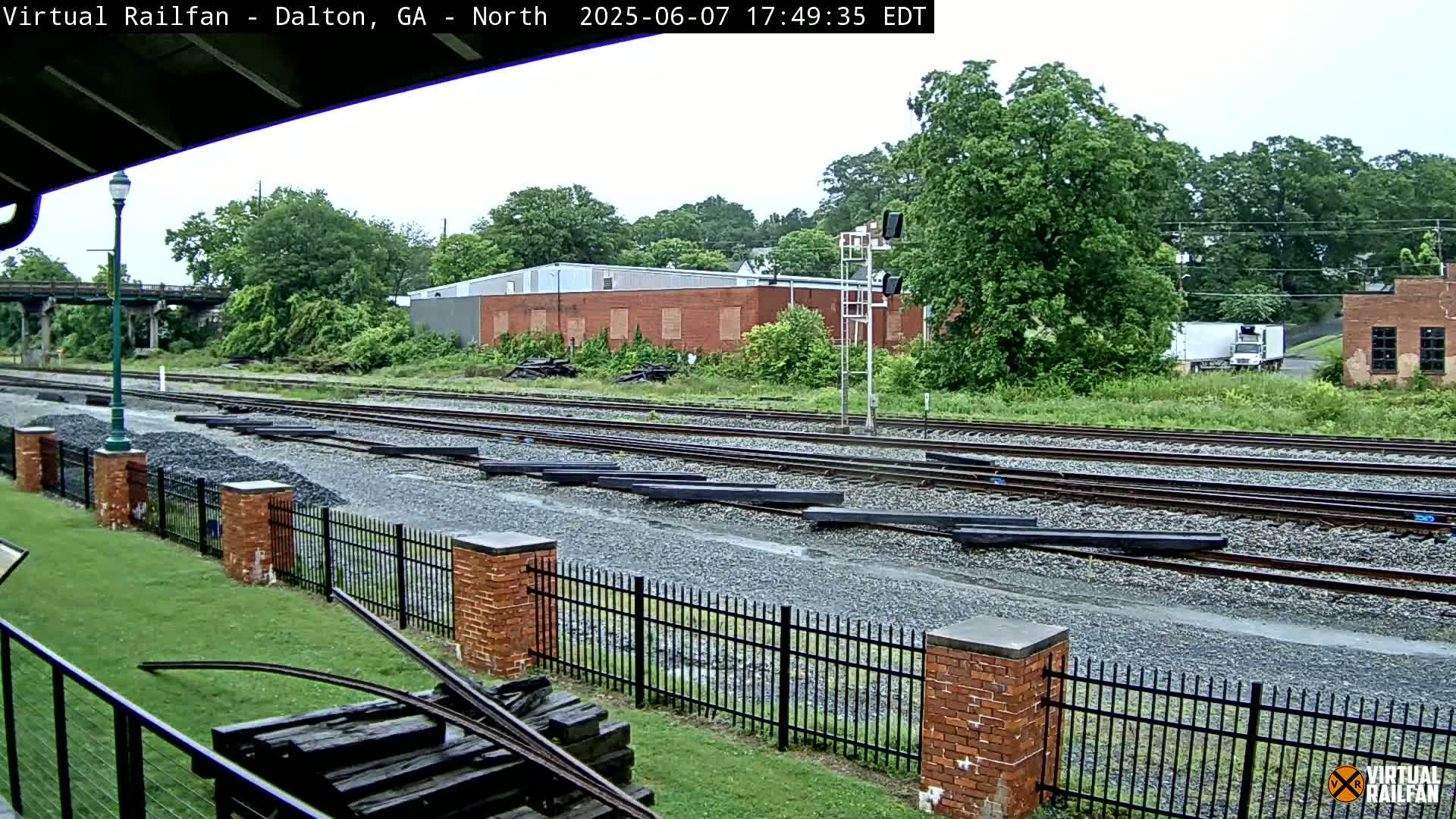 A wet, grassy area next to railroad tracks with a brick fence and several buildings in the background on a cloudy day.