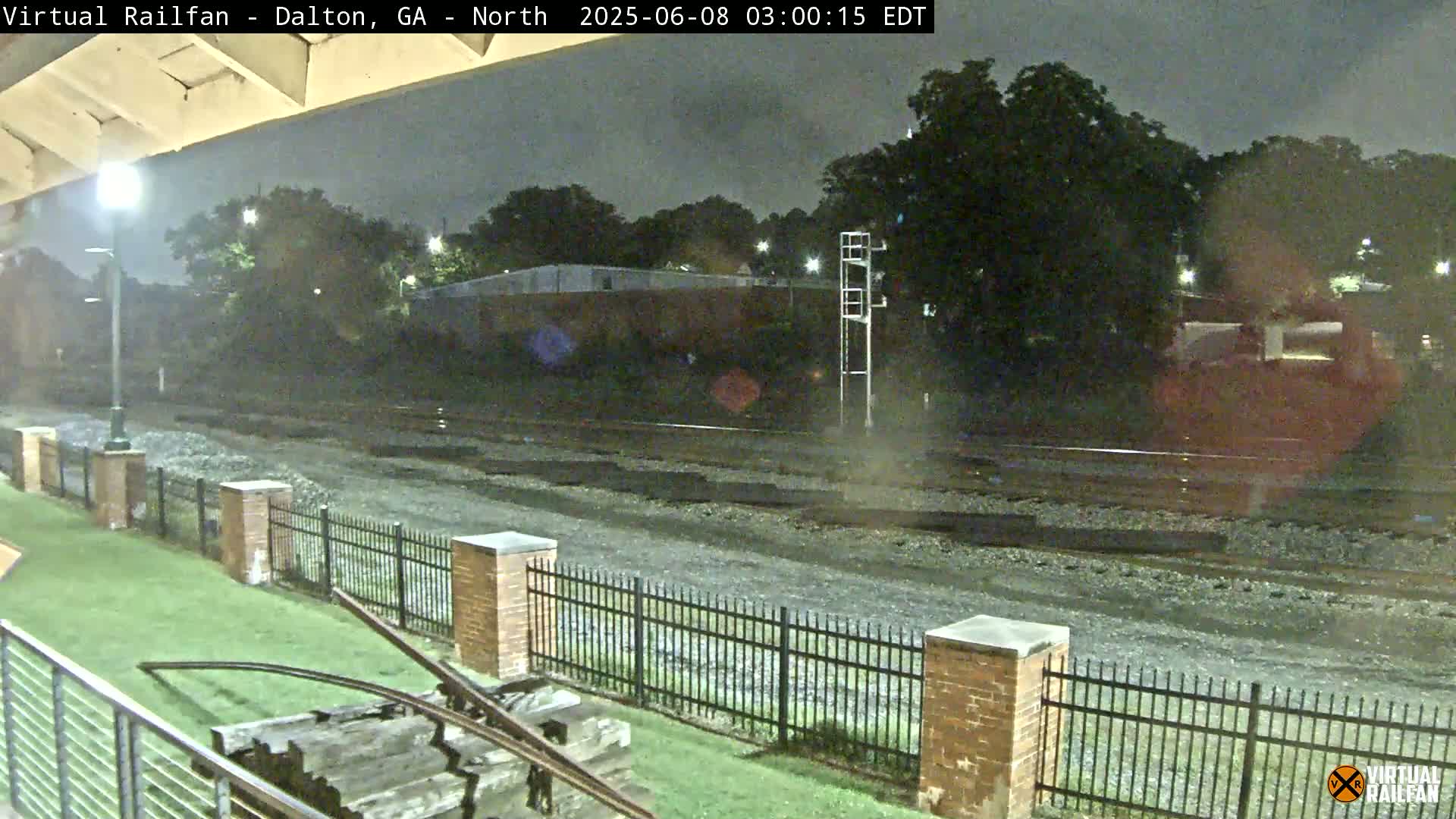 A nighttime view of railroad tracks next to a grassy area enclosed by a brick and metal fence, under a dark sky.