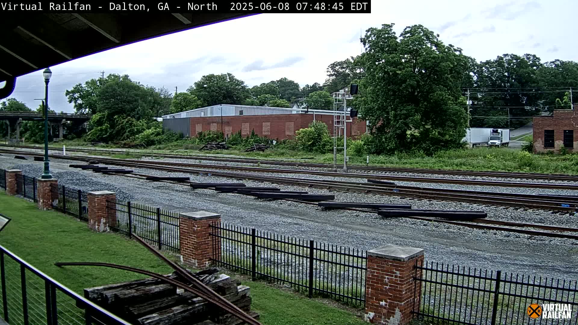 A view of railroad tracks on an overcast day, with several pieces of railway materials stacked nearby and a brick fence separating the tracks from a grassy area.