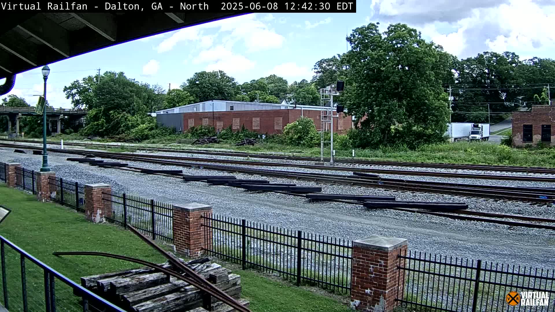A partly cloudy day shows several sets of train tracks with a brick fence and grassy area, alongside a brick building and some trees.