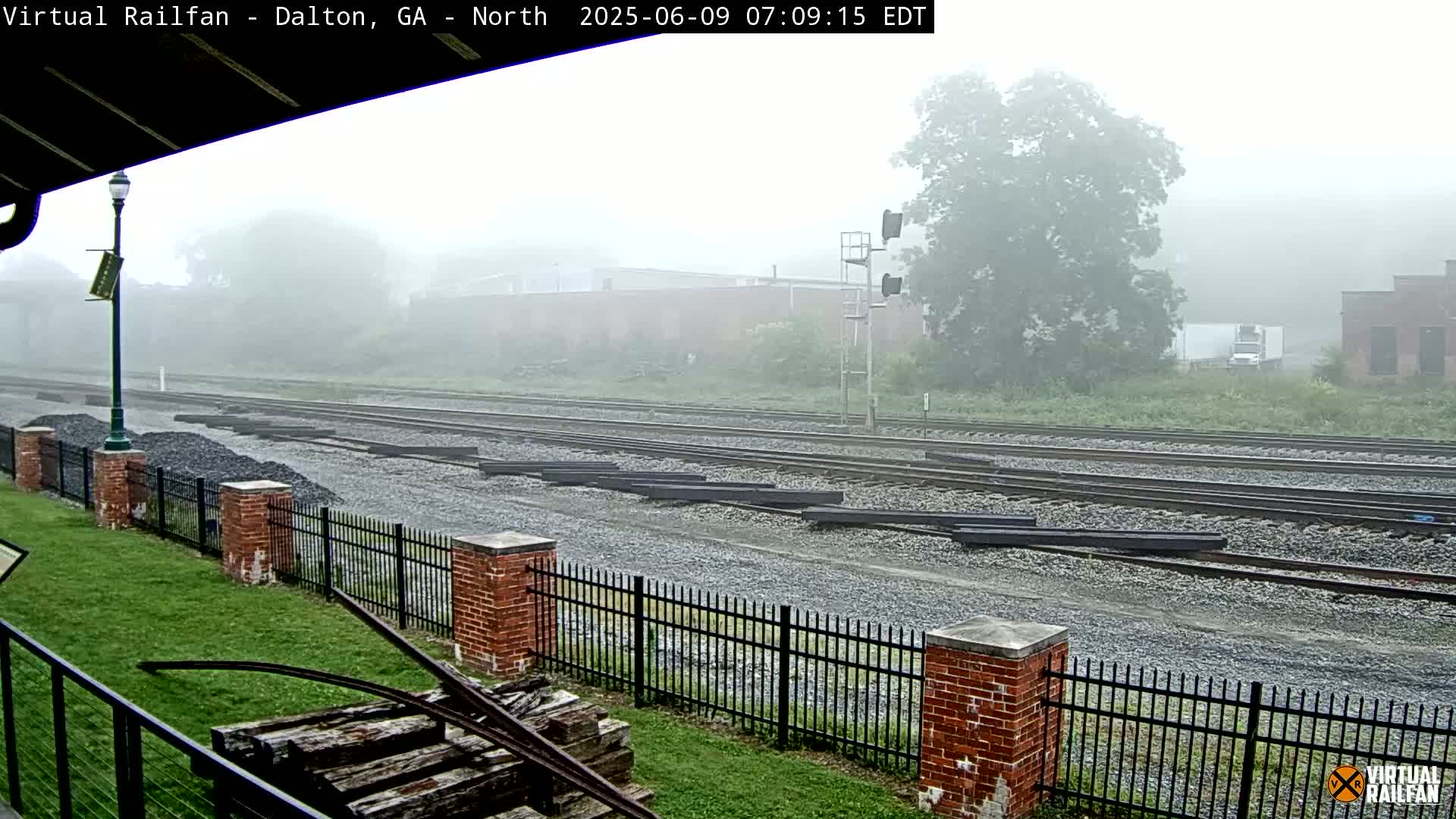 A foggy view of railroad tracks and a brick fence, with a pile of wood and a lamppost visible in the foreground.