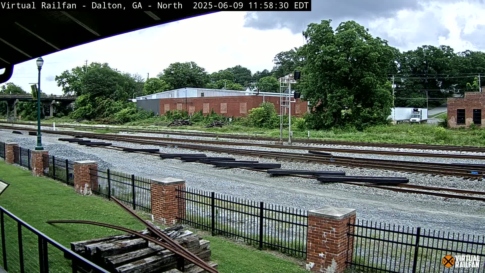Several sets of train tracks stretch into the distance, bordered by a brick fence and lush greenery under a partly cloudy sky.