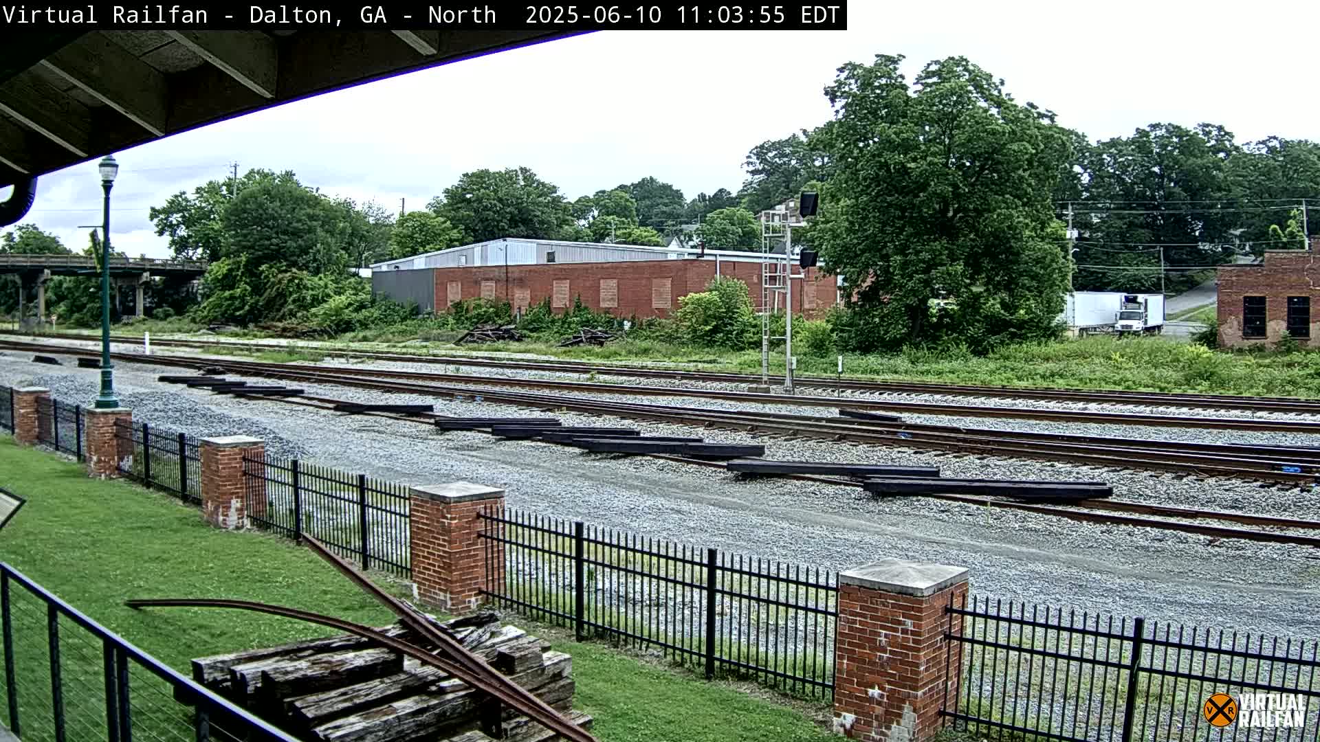 A mostly sunny day shows railroad tracks with several long, dark metal pieces lying on the gravel between the tracks, bordered by a brick fence and lush green grass, with trees and buildings in the background.
