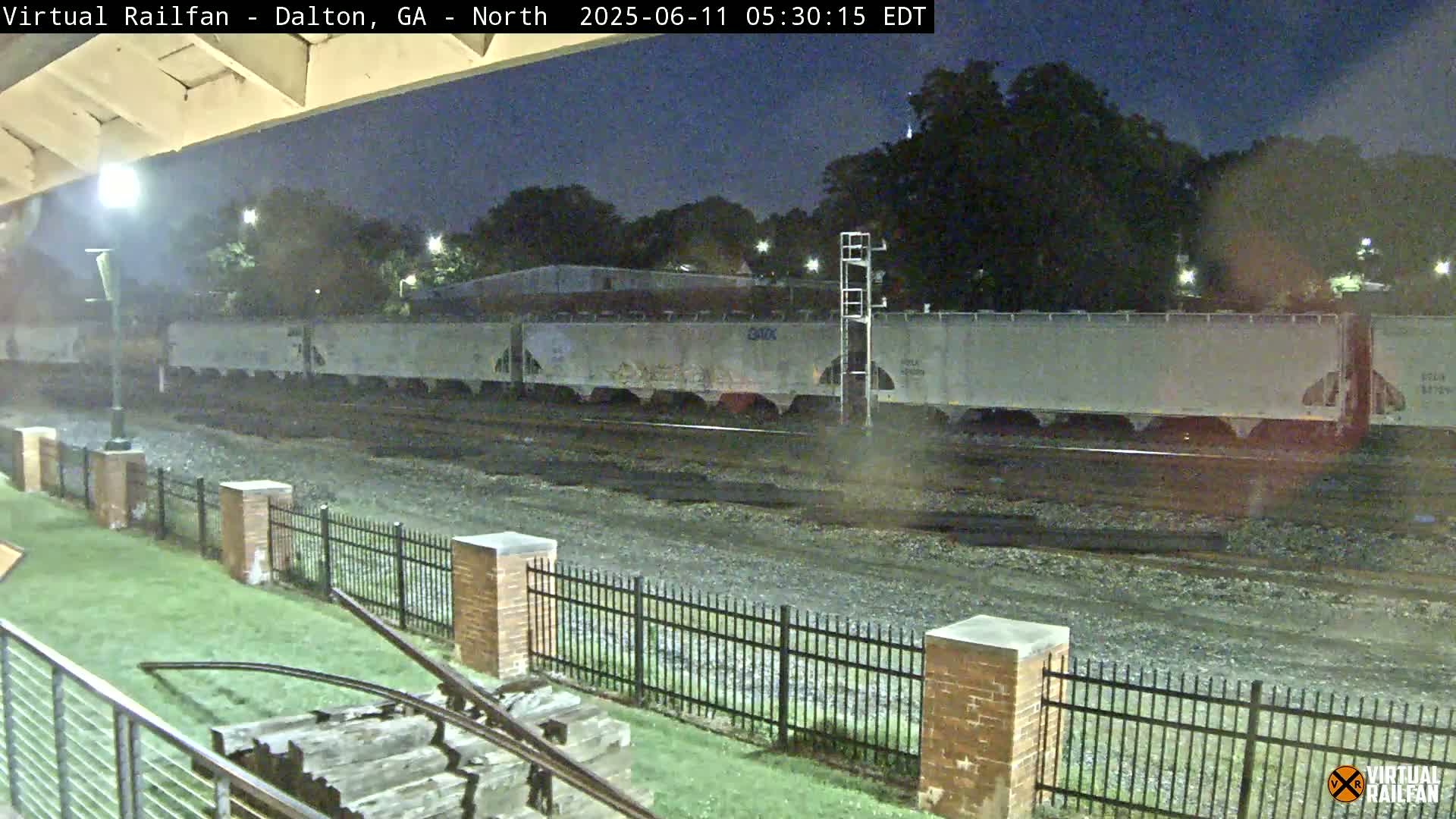 A long freight train sits on railroad tracks at night, behind a short brick fence and grassy area.