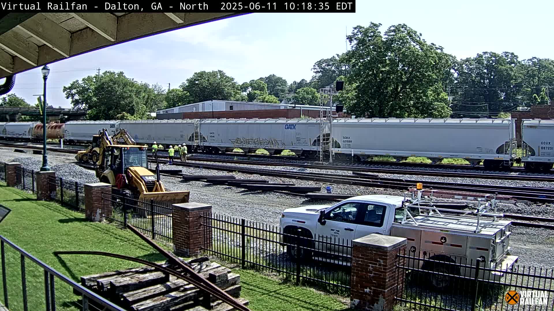 Under a sunny sky, a yellow excavator and several workers are near a long freight train on a set of railroad tracks, with a white pickup truck parked nearby.